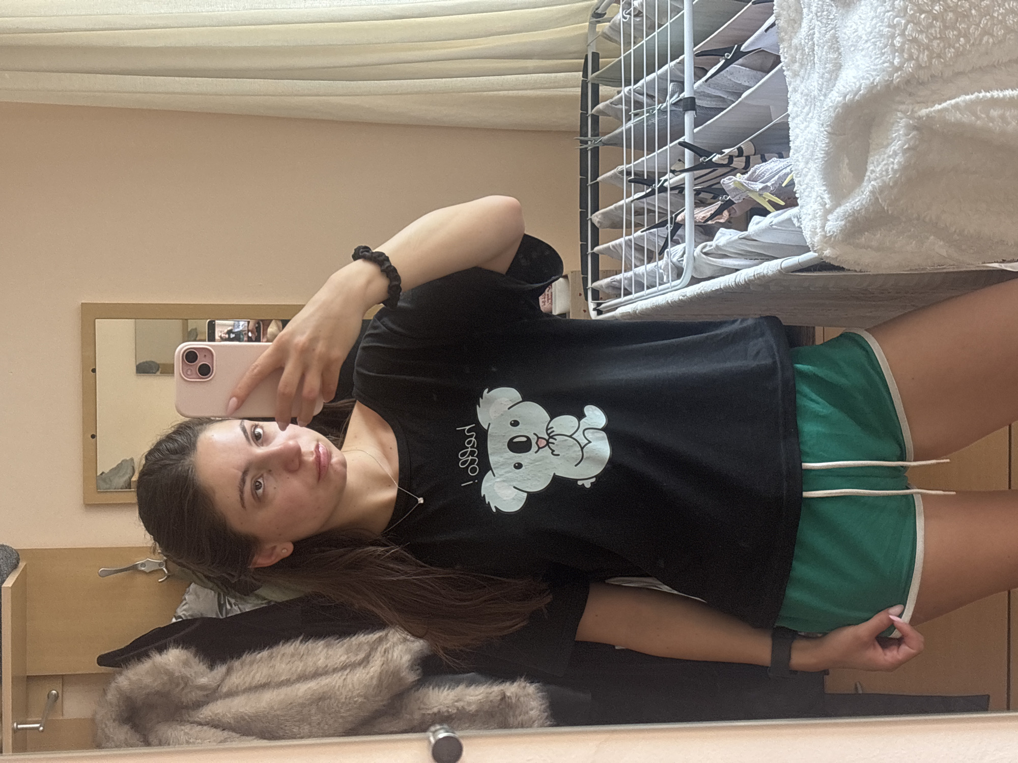 The image shows a young woman taking a mirror selfie in a room. She is wearing a black t-shirt with a cartoon koala graphic and the word "hello!" above it, and green athletic shorts with white trim. She has a black scrunchie on her wrist and a smartwatch on her left wrist. She is holding a pink smartphone with her right hand, angled to capture the reflection. In the background, to the left, is a closet with a fur-lined jacket and other clothing items. To the right, a drying rack filled with laundry stands near a window with sheer curtains. The room appears to be residential, with light-colored walls and wooden furniture. The overall lighting suggests it is daytime. The woman is looking at her reflection, with a neutral expression. The scene is domestic and casual. Varna, Bulgaria does not appear to be depicted in this image.