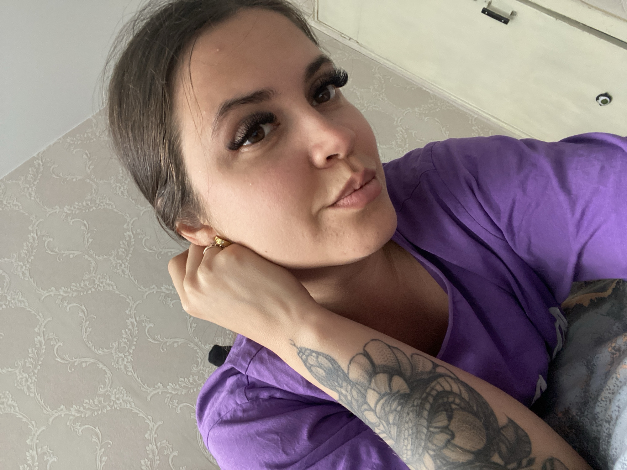 An individual, likely female, is depicted in a close-up, selfie-style photograph taken indoors in Chaikovsky, Russia. She appears to be reclining or lying down, with the camera angled upwards towards her. She has dark hair, styled away from her face, and prominent, voluminous false eyelashes accentuating her brown eyes. Her expression is neutral with a slight pout. She wears a simple purple t-shirt.
Her right hand, adorned with a gold ring and a small gold earring, is visible near her ear, propping up her head. A distinctive feature is a large, intricate black and grey tattoo on her right forearm, depicting what appears to be a snake or a mythical serpent-like creature.
The background features light-colored wallpaper with a decorative, scrolling floral pattern. Above her, a section of a light-colored door or cabinet with a dark latch or handle is visible. The lighting suggests an indoor setting, possibly during the day with diffused light. The overall atmosphere conveyed is casual and relaxed. No visible text is present in the image.