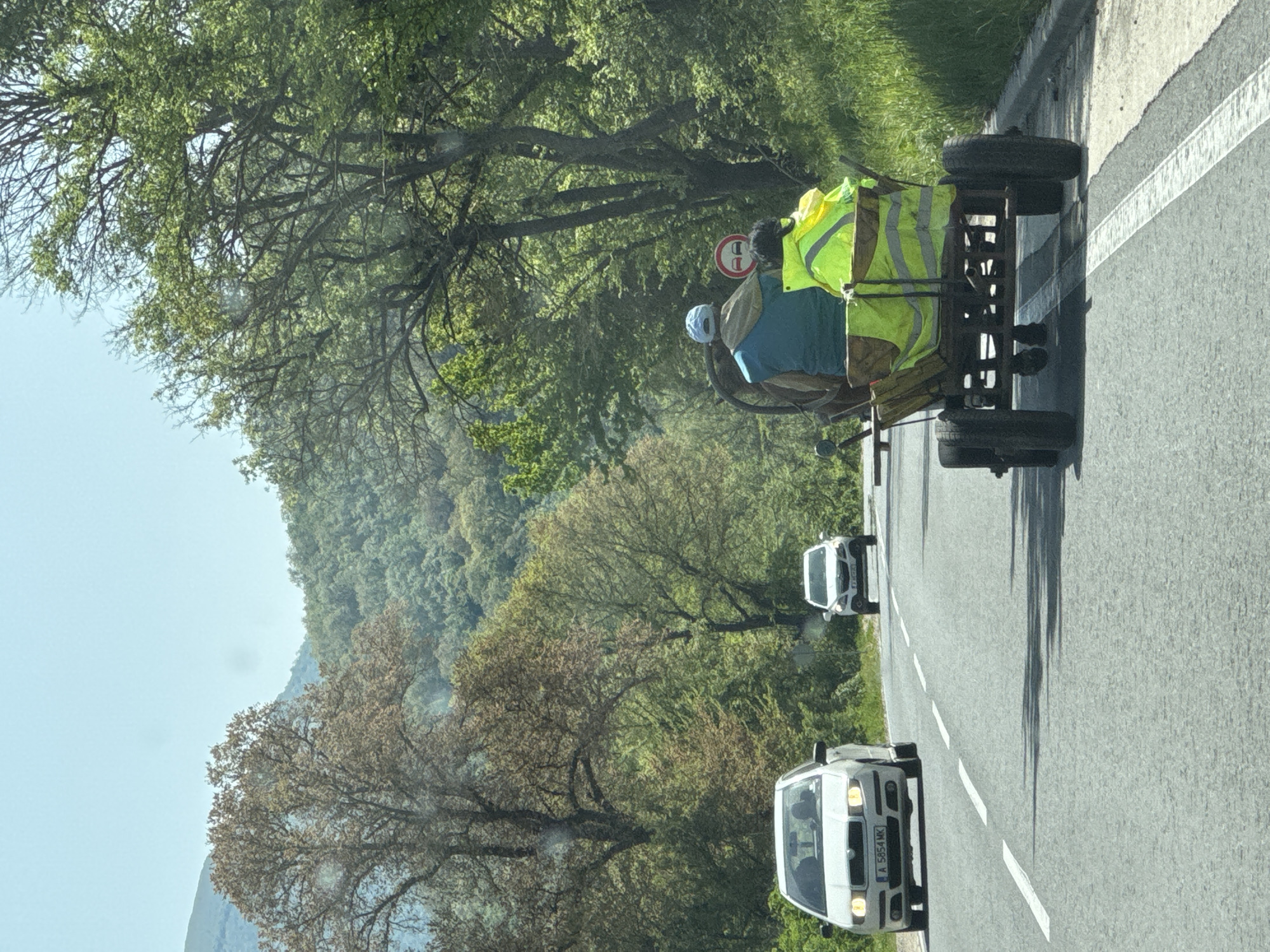 A road in Bilka, Bulgaria, shows a horse-drawn wagon occupied by two individuals wearing safety vests, followed by two cars. The wagon, appearing handmade, occupies the far right lane next to a road divider, suggesting it's sharing the lane with traffic. The cars follow behind, with a white sedan in the foreground. The surrounding environment includes trees and a distant mountain.