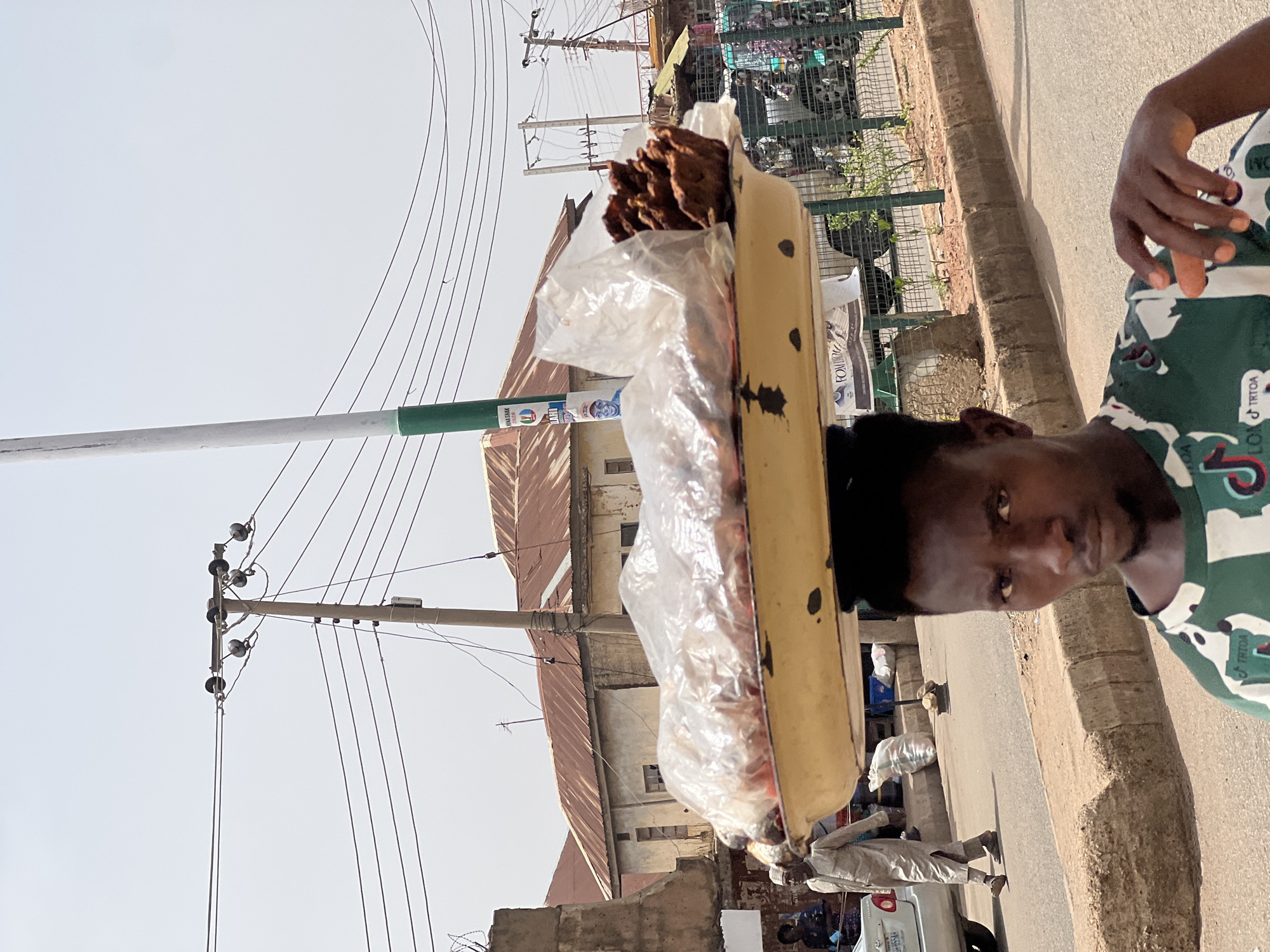 A man in Bukuru, Nigeria, is carrying a tray of food items on his head. The food includes what appears to be meat in plastic bags and loose meat or jerky. The background includes a street, power lines, buildings with corrugated roofs, and other pedestrians.