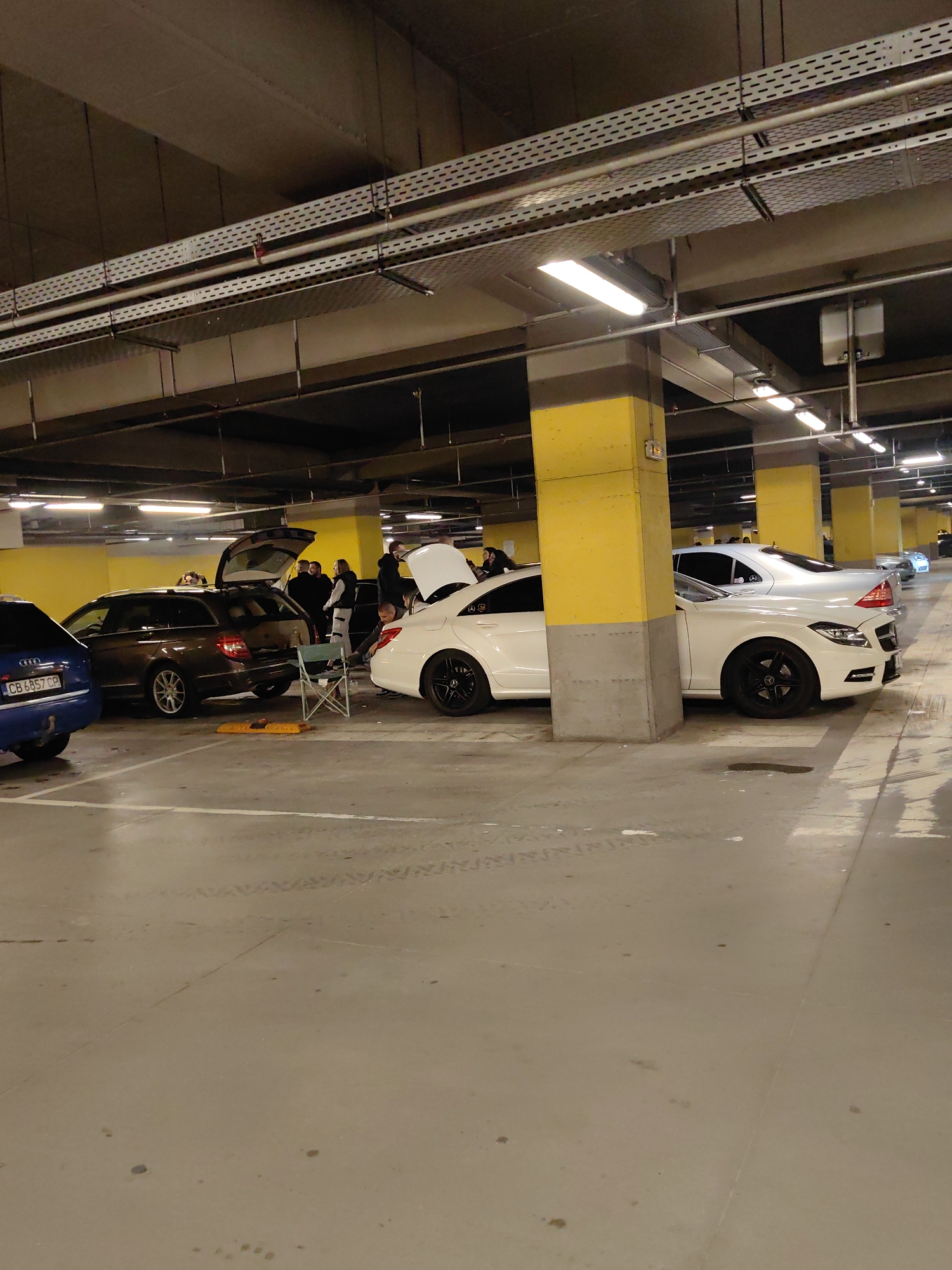 The image depicts an underground parking garage in Sofia, Bulgaria, evidenced by the license plates on the vehicles. The garage has yellow-painted support columns and a high ceiling with exposed pipes and lighting fixtures. Several cars are parked, including a blue Audi, a brown Mercedes-Benz station wagon with its trunk open, and at least two white Mercedes-Benz CLS cars with modified features (spoilers, black rims), also with open trunks. A group of people surrounds the brown Mercedes, and one person sits on a folding chair near the car. The floor is marked with parking space lines.
