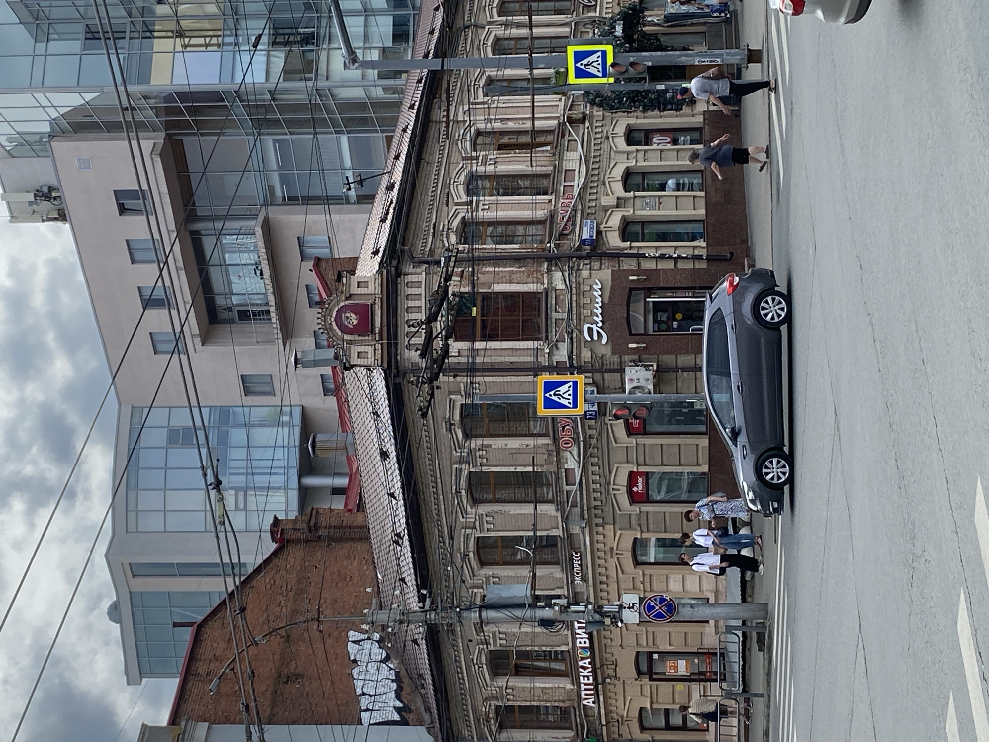 The image shows a street scene in a city. In the foreground, a gray hatchback car is driving across a crosswalk. To the right, a silver sedan is partially visible, also on the road. On the sidewalk to the left, a group of three people, two men and a woman, are standing near a traffic light and a pedestrian crossing sign. Another woman is walking away from the camera on the sidewalk.
The main building in the midground is an older, ornate structure with light-colored brickwork and a red tile roof. Several windows with dark frames are visible. Signs on the building include "АПТЕКА ВИТ" (Pharmacy VIT) and "Эспрессо" (Espresso). Another sign reads "Обувь" (Shoes). A modern glass-fronted building is visible behind and to the right of the older building, reflecting the sky and surrounding structures.
The environment appears to be a city intersection with traffic lights, street signs, and power lines overhead. The sky is overcast with gray clouds.