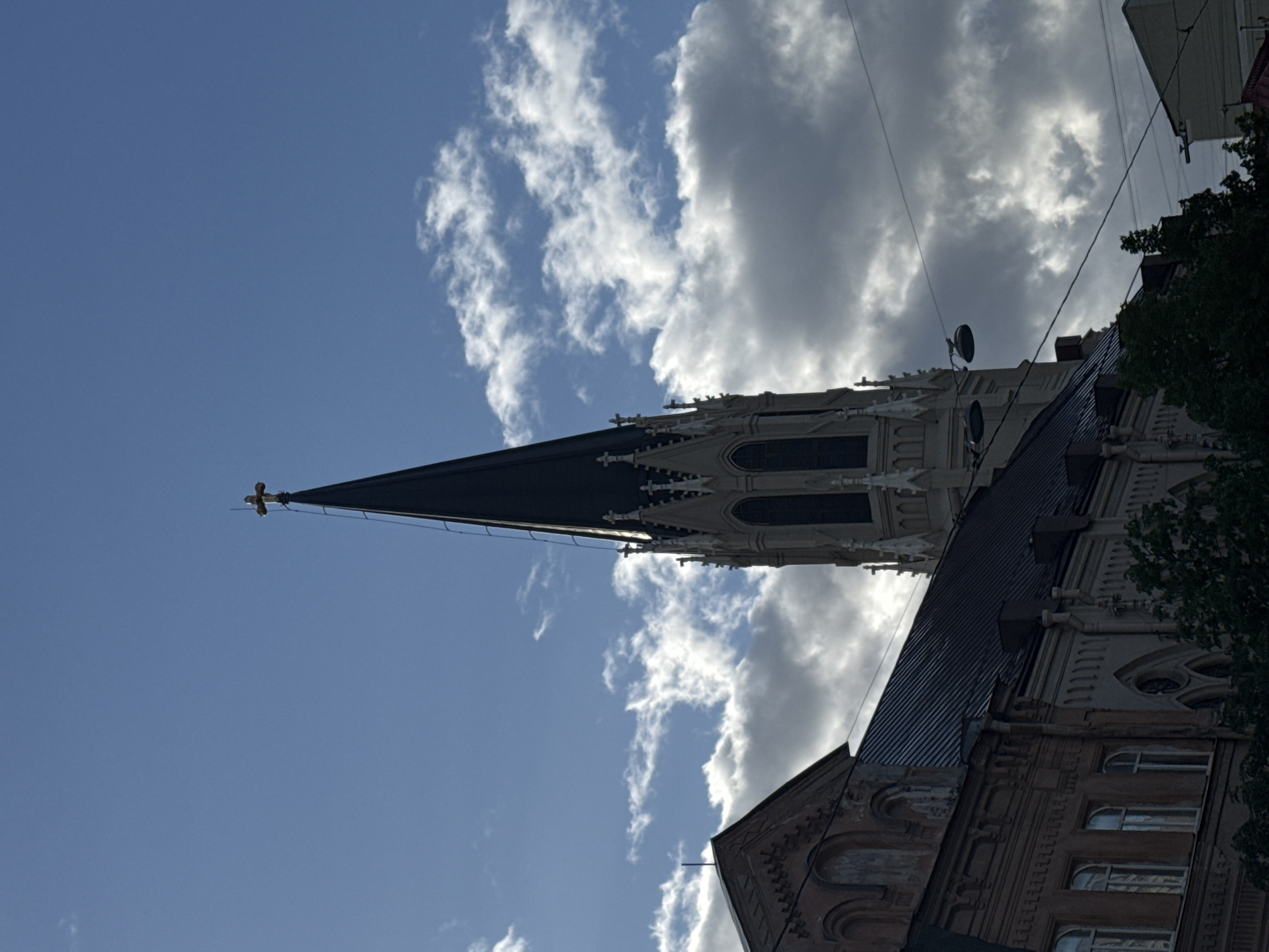 The image depicts the steeple and part of the facade of Saint Peter's Church in Saint Petersburg, Russia, against a cloudy sky. The steeple is black with a gold cross at its apex. The church's facade is light-colored stone with Gothic architectural details. A section of a building with brick details is visible in the lower right corner. Electrical wires cross the sky. The image is taken from a low angle, looking up at the church.