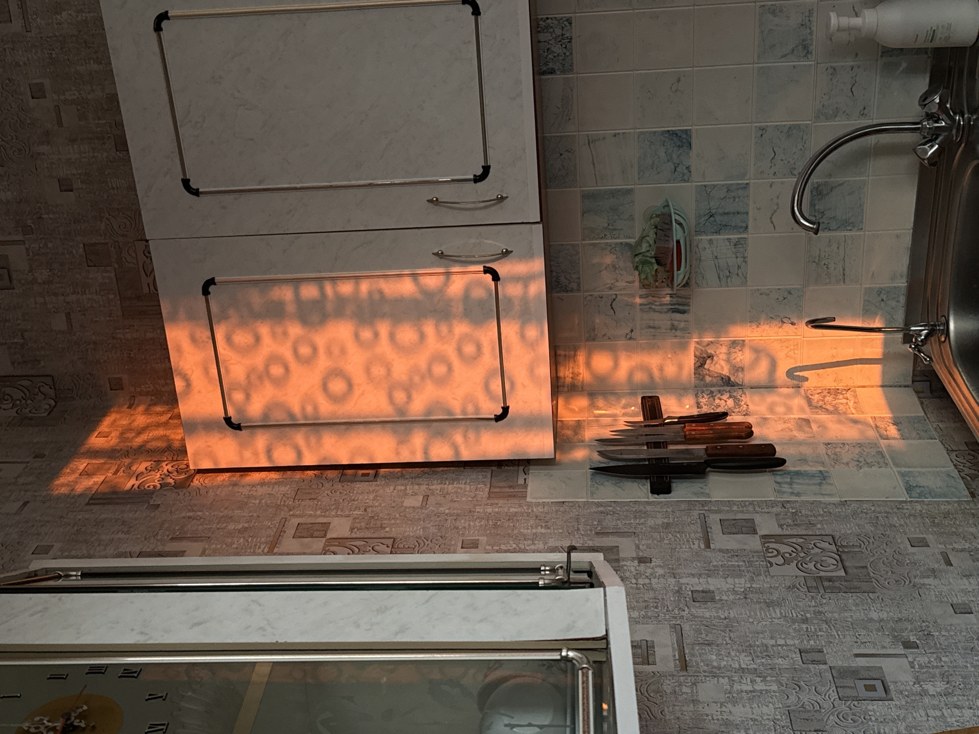 The image depicts a section of a kitchen interior. Two kitchen cabinets are visible, stacked one on top of the other. To the right is a sink with a faucet and soap dispenser. On the tiled countertop are several knives in a knife holder. The floor tiles continue the pattern from the countertop. Part of a stove with a glass door is visible in the foreground. A warm light is cast across the scene, illuminating the objects and surfaces. Based on image context clues like typical kitchen tiling and cabinets, and architectural style common in older residential buildings, a location in Saint Petersburg, Russia, is speculative but plausible. No people are visible.
