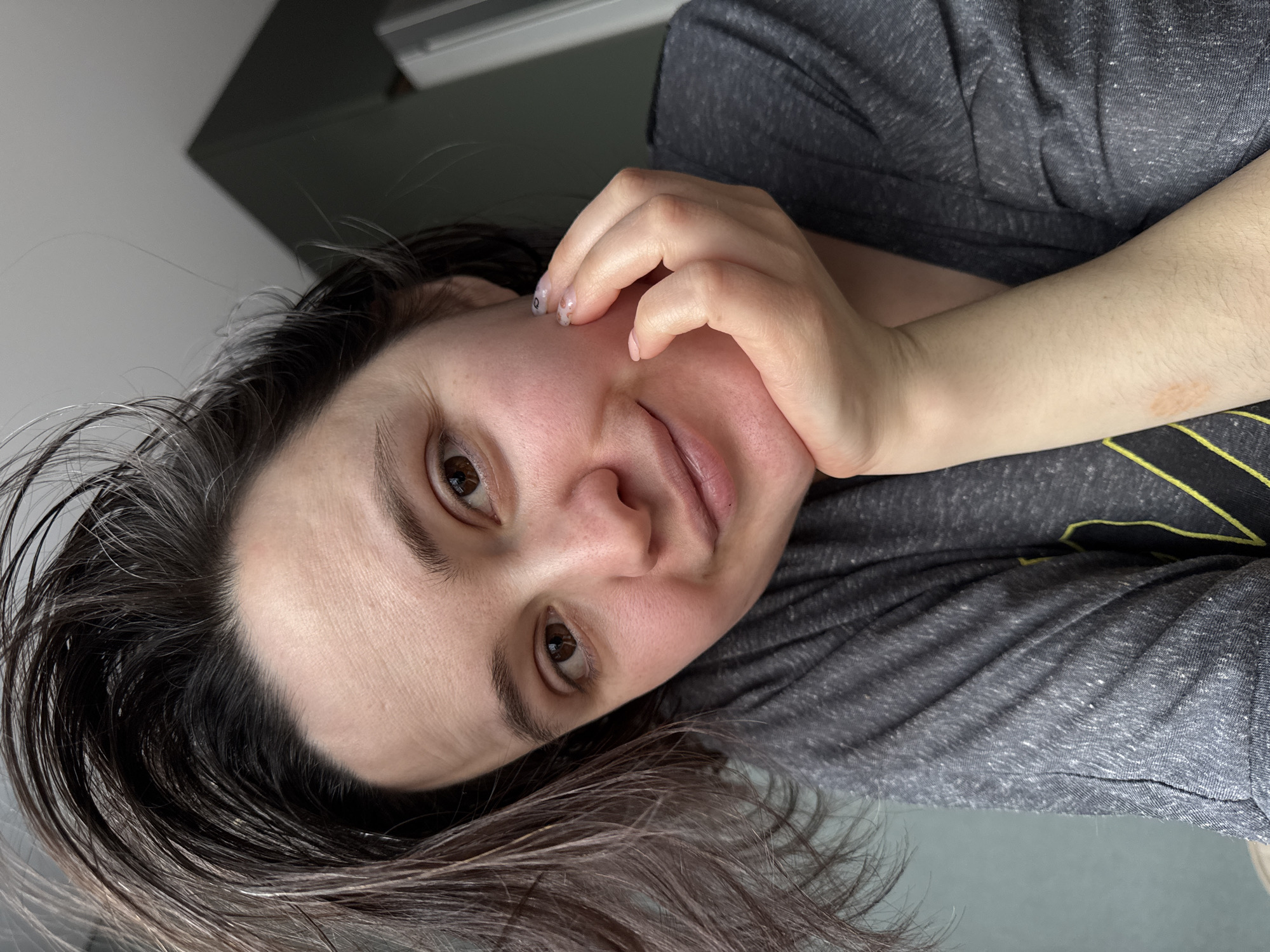 A woman is centered in the image, facing the camera with her head tilted to the left. She is wearing a gray shirt. Her right hand is held up to her cheek. Her hair is dark and partially wet, with a slight reddish tint at the ends. The background shows a blurry gray surface. There is no information suggesting the location of the picture is in Izhevsk, Russia.