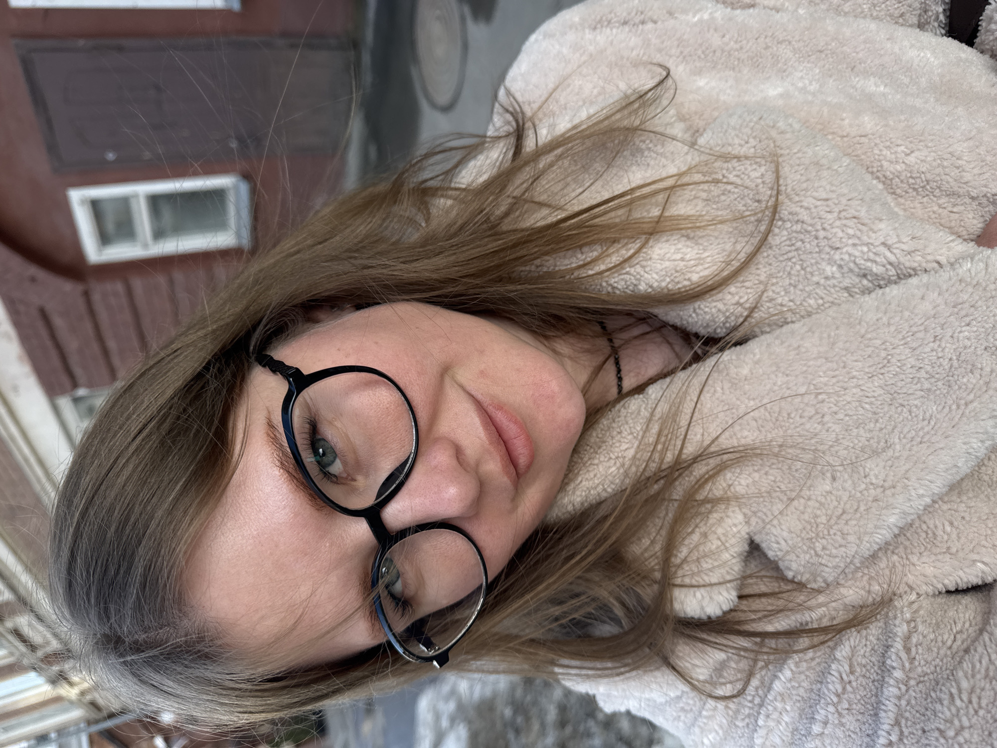 A woman with long brown hair and round black glasses is pictured in an outdoor urban setting. She wears a fluffy, light-colored jacket. Behind her, a red brick building with a white-framed window can be seen. A section of paved ground is also visible, suggesting a street or sidewalk. The photo's location is Perm, Russia.