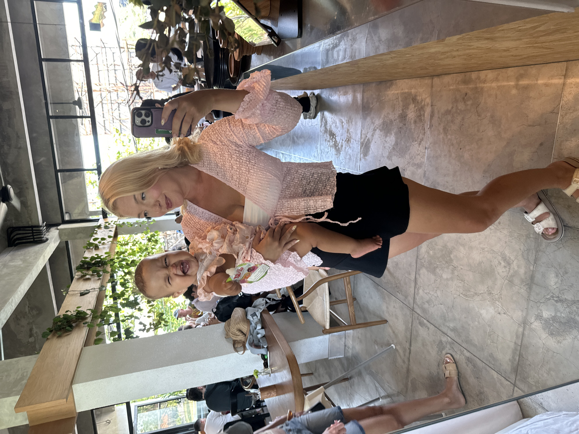 A blonde woman in a light pink ruffled top and black shorts is taking a mirror selfie. She is holding a baby wearing a pink ruffled onesie. The baby is smiling widely, with its eyes closed and mouth open. The scene appears to be indoors, possibly a cafe or restaurant, with concrete floors and a rustic wooden counter. There are other people visible in the background, some seated at tables. The lighting is bright and natural, suggesting daytime. The location is North Kuta, Indonesia.