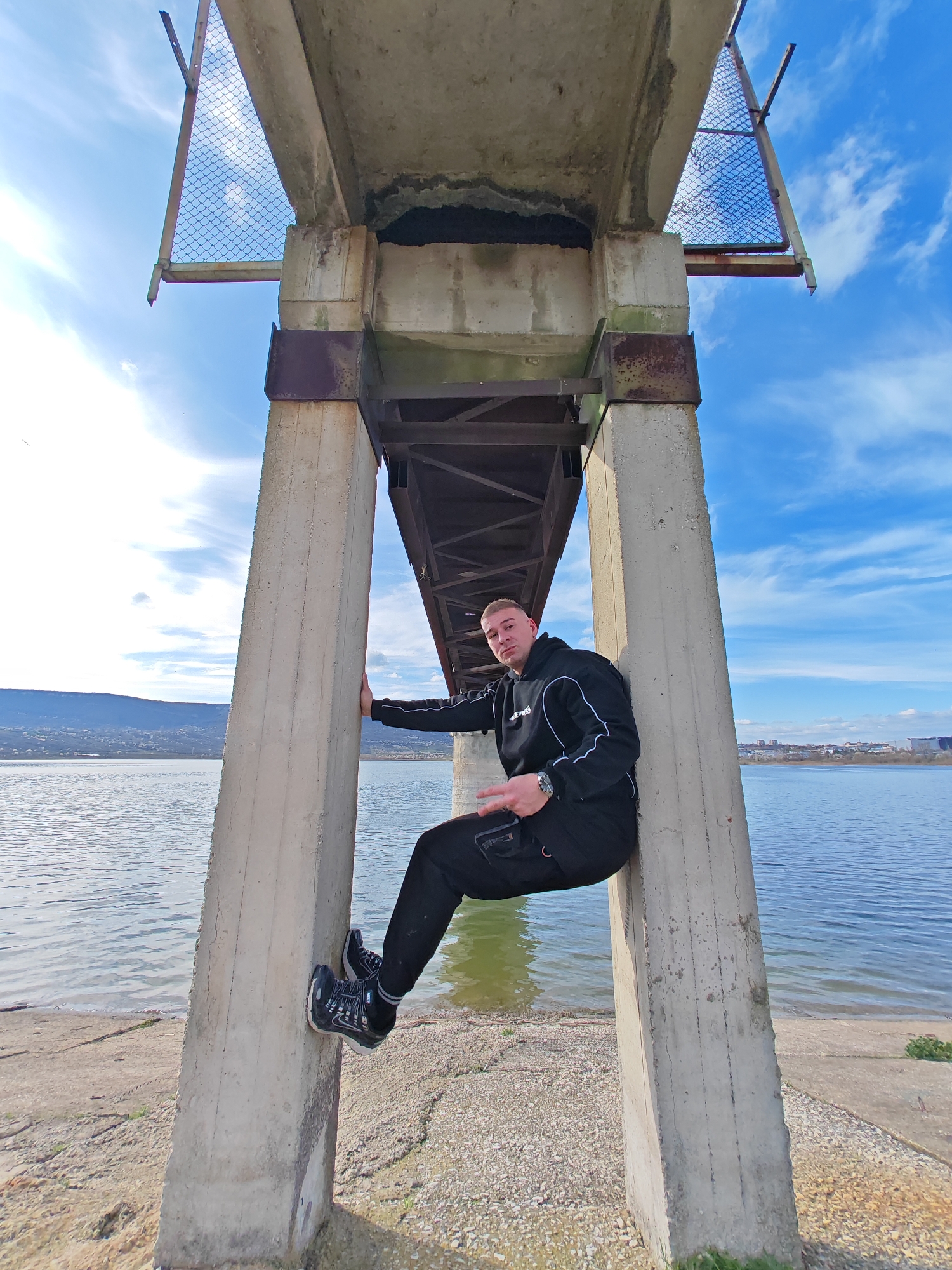 A full shot shows a man sitting on a concrete support structure of a bridge. The man is fair-skinned with short blond hair and is wearing a black tracksuit and dark sneakers. He is sitting with one hand on the support and the other holding a dark object. His legs are bent, and his feet are resting on the lower section of the support. The scene is set under the bridge, looking outward towards a large body of water. The sky is blue with scattered clouds. In the distance, there are hills and a cityscape. The ground beneath the bridge appears to be a mixture of gravel and concrete. This bridge is located in the city of Shumen, Bulgaria.