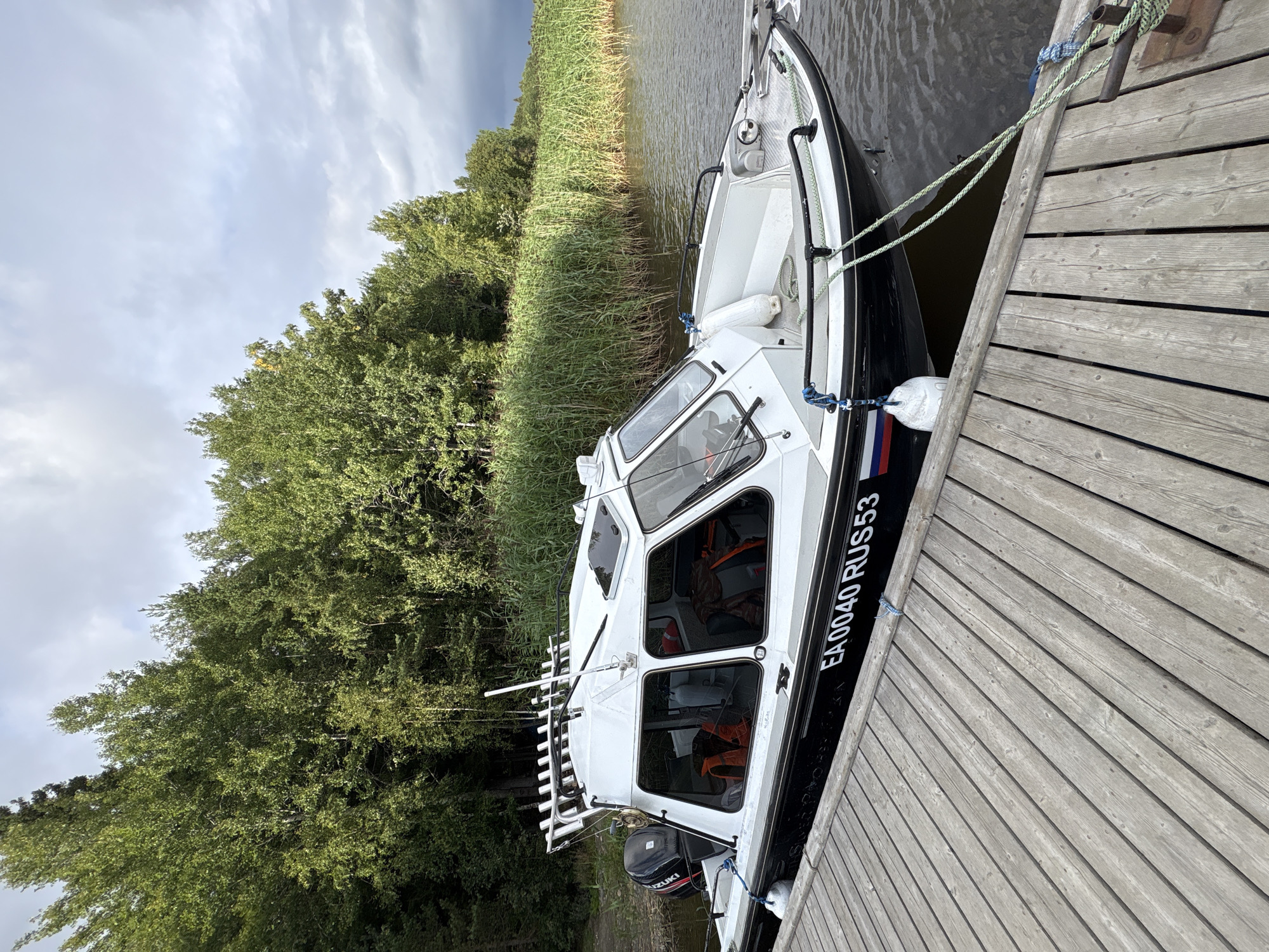 A white and black motorboat is docked at a wooden pier. The boat has a cabin with windows and a metal railing. An outboard motor is attached to the stern. The boat's hull is marked with "EA0040 RUS53" and a Russian flag. Orange life vests are visible inside the cabin. The pier is made of weathered wooden planks. Behind the boat, there is a body of water and a dense line of tall green reeds. Beyond the reeds, a forest of green trees extends towards a cloudy sky. The scene appears to be a waterfront area in Rautalakhti, Russia.