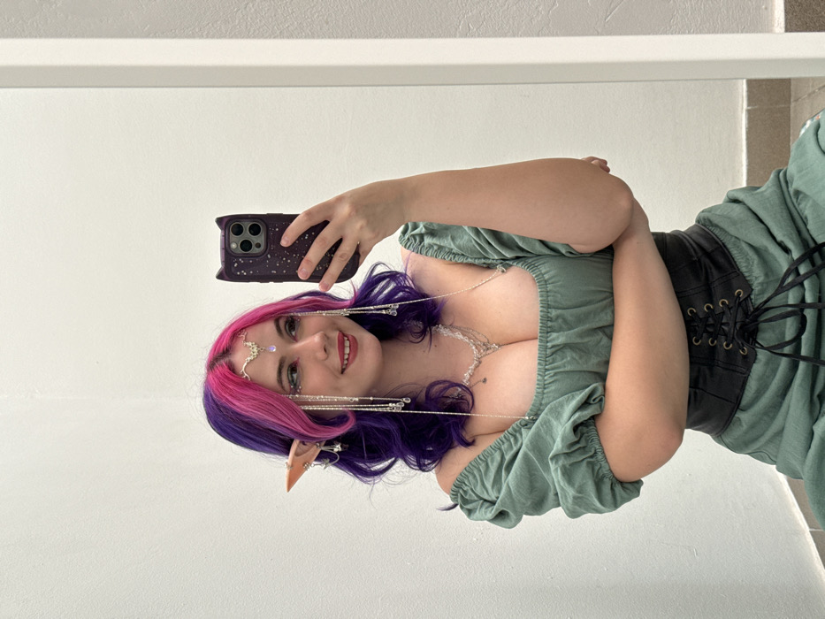 A woman with purple and pink hair, pointed elf ears, and a beaded headpiece takes a selfie. She wears a low-cut green blouse with puffy sleeves and a black corset. She holds a phone with a dark, star-patterned case. The background is a white wall with a horizontal white molding. She smiles broadly, showing red lipstick.