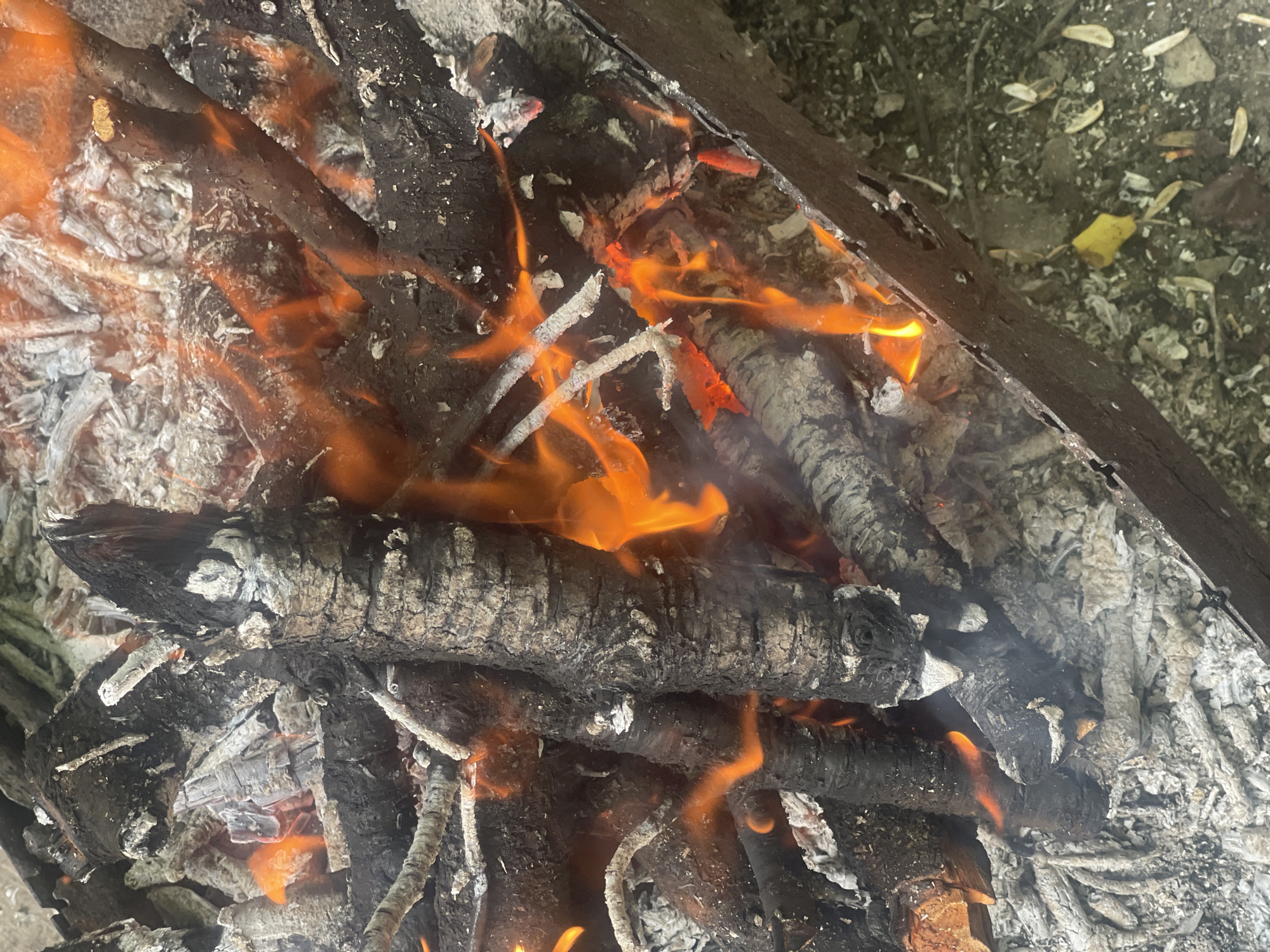 The image shows a close-up view of a bonfire in a metal fire pit, situated outdoors on the ground. The fire pit is partially filled with burning logs, ashes, and embers. Flames in shades of orange and yellow are actively burning, illuminating the scene. The logs are a mixture of dark, charred wood and lighter, ash-covered pieces. The surrounding ground appears to be dirt or gravel, with some scattered debris. The location is identified as Ohuivka, Ukraine. There are no people or animals visible in the image. The focus is entirely on the fire and its immediate surroundings.