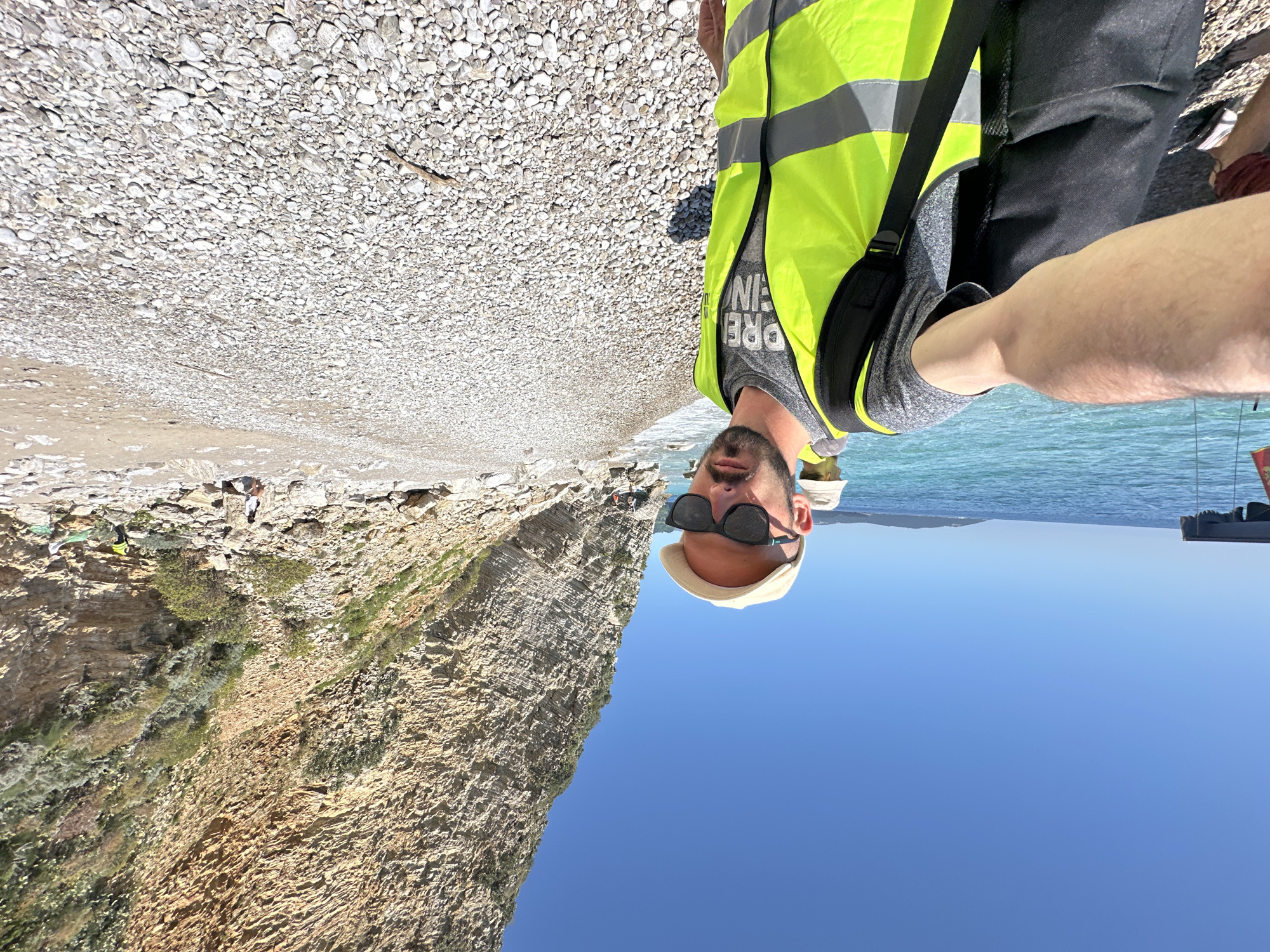 A man wearing a yellow reflective vest, gray shirt, black pants, and a tan baseball cap is taking a selfie on a beach. He also wears sunglasses. He stands on a pebble beach, with the Adriatic Sea in the background. To the left of the frame is a cliff face. The sky is clear and blue. The photo appears to be taken on Mogren Beach near Budva, Montenegro, based on the landscape. The angle of the photograph is upside down.