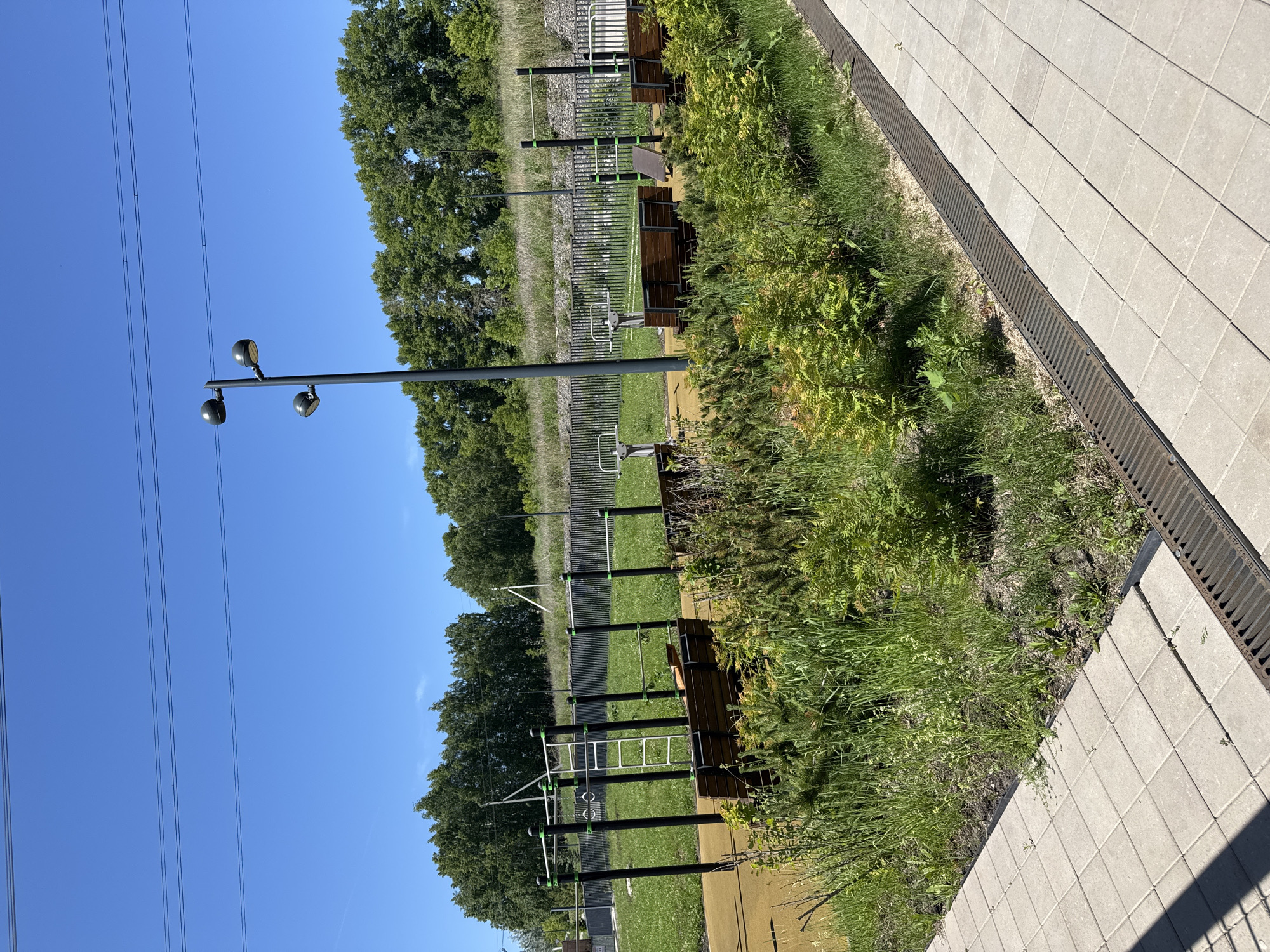 This image shows an outdoor sports and recreation area with exercise equipment and benches, set against a backdrop of trees and a clear blue sky. The area is paved with light-colored square tiles, and a metal grate runs along the edge of the pavement. In the foreground, there are low-lying green bushes and tall grass.
The sports equipment includes horizontal bars, parallel bars, gymnastic rings, and a ladder, all constructed with black metal poles and green accents. Several wooden benches are placed near the equipment, some with slatted backs and seats. In the background, a line of lush green trees borders the area. A tall lamp post with three lights stands prominently on the right side of the frame. Power lines are visible crossing the sky.
The image appears to be taken during the daytime, under bright sunlight, suggesting a warm day. Based on the context of the surroundings, this location is likely in the city of Krasnoyarsk, Russia, possibly in a park or a dedicated public space designed for outdoor fitness and leisure. There are no people visible in the image.