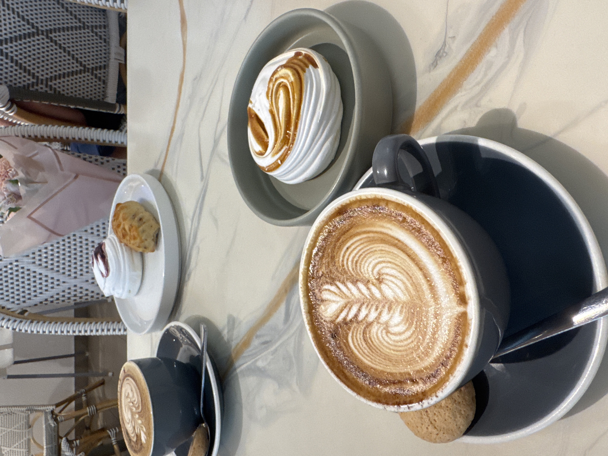 This is a close-up, eye-level shot of two cups of coffee with latte art and two meringues with caramel drizzle, placed on a marble-patterned table. The scene is likely in a cafe or restaurant in Denpasar, Indonesia.
In the foreground, a dark grey coffee cup with intricate latte art, a small cookie, and a spoon sits on a matching saucer. To its right, a grey bowl holds a white meringue with a swirling caramel topping.
In the background, another dark grey coffee cup is visible on a saucer. Behind it, on a white plate, are two meringues with a dollop of red jam on one and a small pastry on the other. Behind these treats, a bouquet of pink and white flowers is partially visible, held by someone seated at the table. The chairs at the table are woven with a patterned design. The overall impression is one of a pleasant cafe setting.