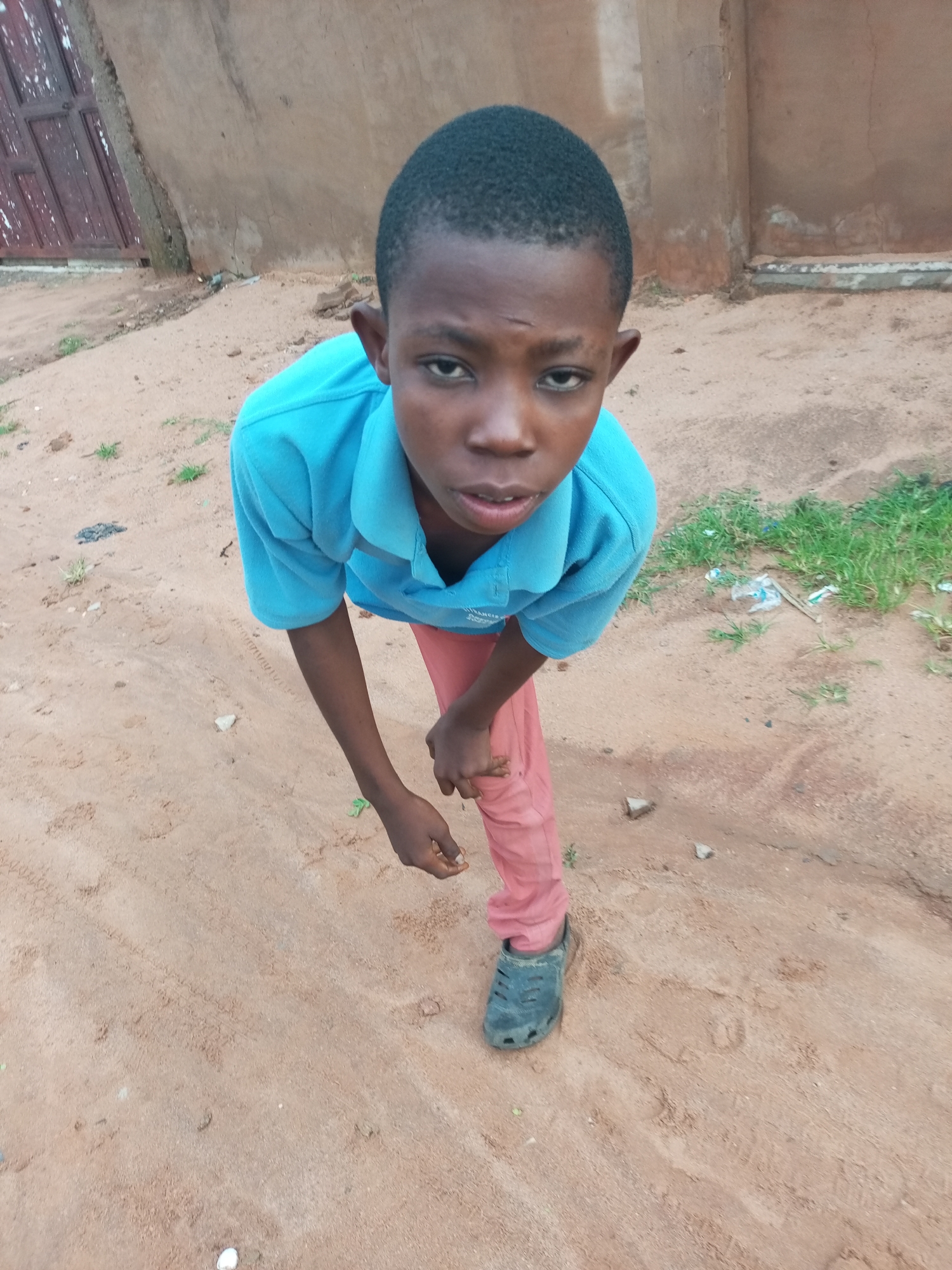 A young boy, appearing to be of Nigerian descent, is captured in a medium shot, looking directly at the camera with a slightly concerned expression. He is wearing a bright blue polo shirt and reddish-pink pants. His left hand is held near his waist, and his right hand is resting on his right leg. The boy is standing on a dirt-covered ground, which appears to be a road or pathway, with scattered green grass and some debris visible in the background. Behind him, a textured wall, possibly made of concrete or stucco, runs horizontally, with a wooden door to the left. The scene suggests an outdoor setting in a potentially rural or less developed area of Tempure, Nigeria, given the earthy environment and simple structures. The lighting indicates it may be daytime, possibly overcast, as there are no strong shadows.