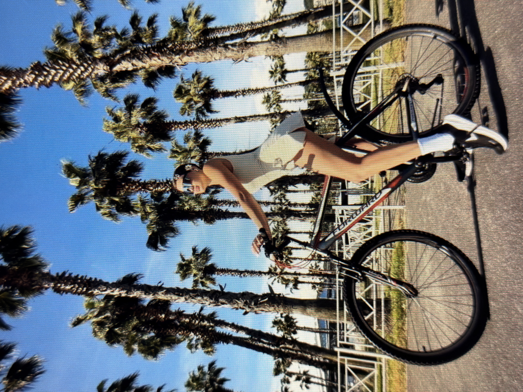 An outdoor, eye-level, full shot captures a young woman on a bicycle in Sirius, Russia, with a backdrop of palm trees against a clear blue sky. The woman, wearing a white tennis dress and sunglasses, is positioned mid-pedal. Her right foot is on the pedal, and her left foot is extended behind the bicycle's crank. The bicycle is a mountain bike with black tires and a black frame accented with red. She is looking to her left, with her hair pulled back and a slight smile on her face. The environment appears to be a paved walkway or road, with a white railing and lush green grass visible behind it, bordering a line of numerous palm trees. The lighting suggests it is daytime, with clear shadows cast by the woman and the bicycle.