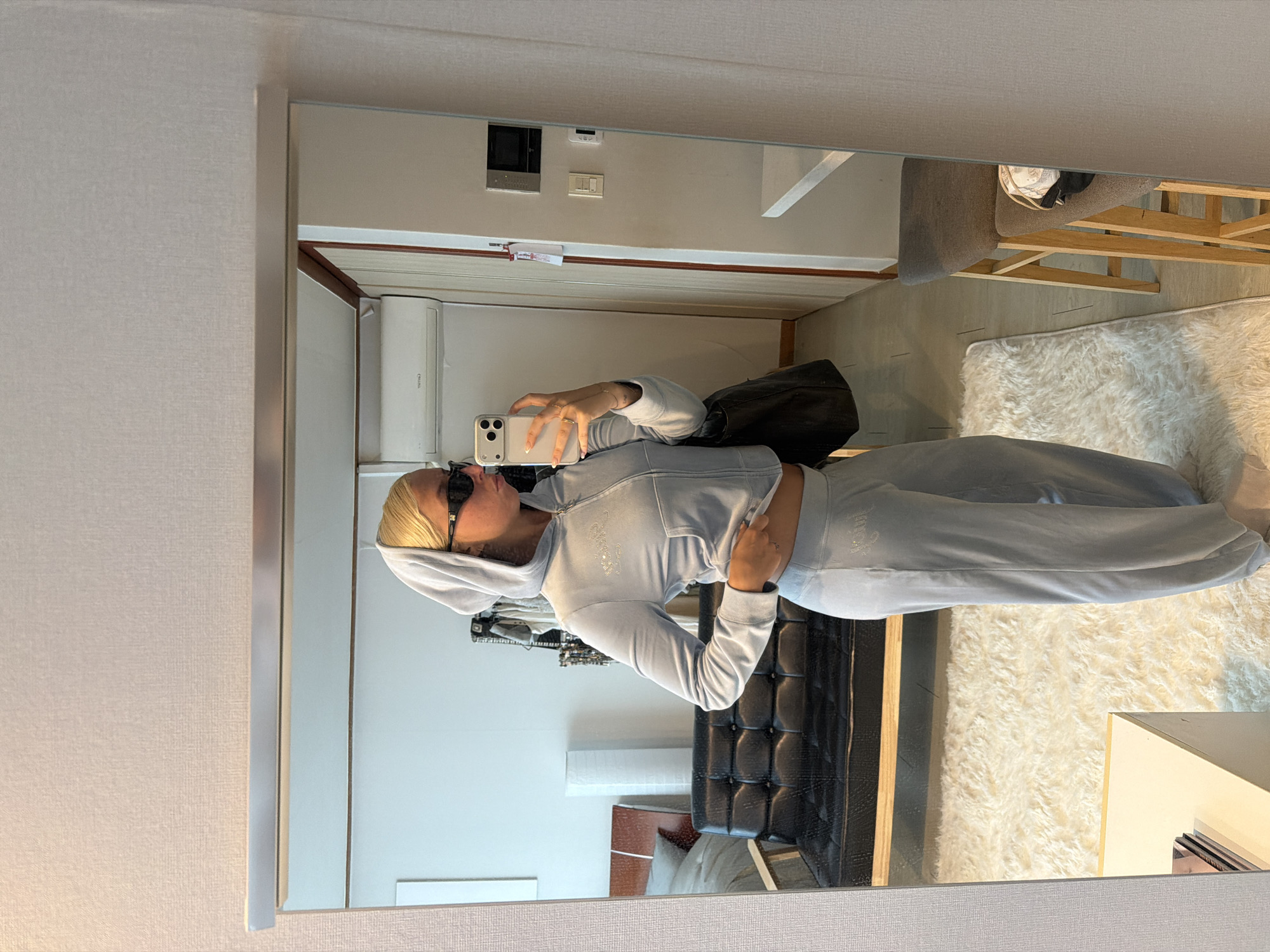 A person with blonde hair and black sunglasses is captured in a mirror selfie, posing casually. They are wearing a light blue or light grey velour tracksuit, consisting of a hooded jacket and matching pants. The hoodie features a glittery text, likely "Juicy," on the left chest area. The individual holds a light-colored smartphone with their right hand to take the photo, while their left hand rests on their stomach, slightly lifting the hoodie. They wear several rings on their fingers, and their nails have white polish.
The reflection in the mirror reveals an indoor room, suggesting an apartment or hotel room in Seoul, South Korea. In the background, a black tufted headboard or a similar piece of furniture is visible. The floor is partly covered by a fluffy, light-colored rug, and a bed frame with wooden slats supporting a mattress can be seen beneath it. A grey upholstered chair with wooden legs is also partially visible.
Higher up on the wall, an air conditioning unit is installed. Below it, a dark rectangular object (possibly a control panel or small screen) and two light switches are mounted. A small white paper or sign is taped to the wall below these fixtures. The room appears to be illuminated by indoor artificial lighting.