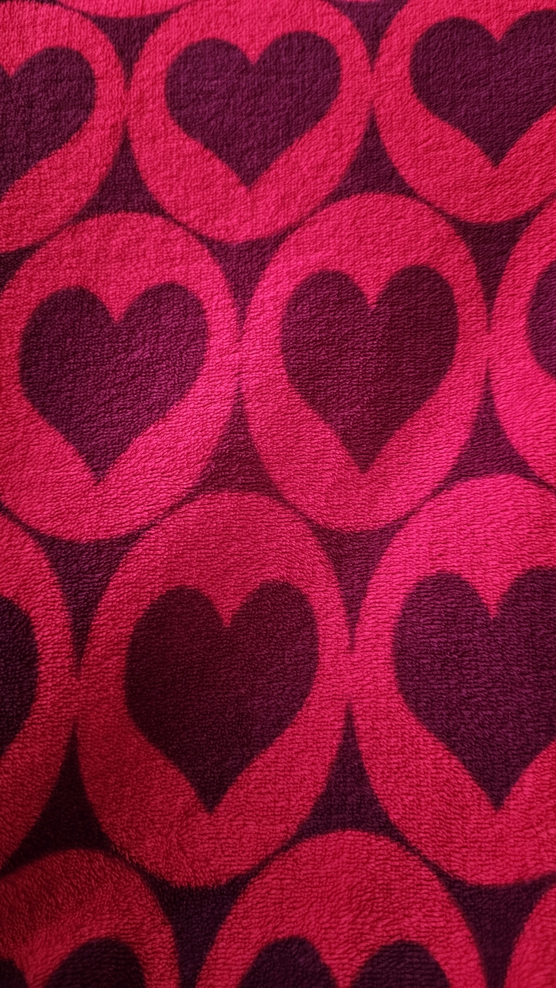 The image is a close-up, full-frame photograph of a textured fabric with a repeating pattern of stylized hearts. The hearts are a bright, saturated pink, outlined by a deeper, darker magenta or deep purple background. The texture appears fuzzy or plush, likely from a terrycloth or similar material. The hearts are arranged in a staggered grid, with each heart contained within an implied circle, and the circles overlapping slightly. The lighting is somewhat harsh, creating bright highlights on the pink surfaces and casting subtle shadows that emphasize the texture and depth of the fabric. There are no people, actions, or environmental elements visible in the image, and no discernible location in Rostov-on-Don, Russia. The photograph solely depicts the textile pattern.