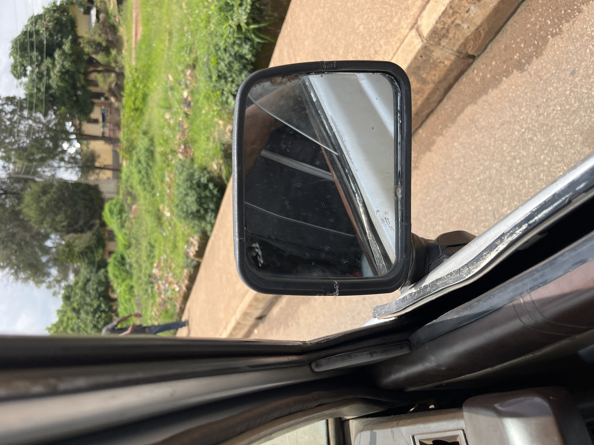 This image captures a perspective from inside a vehicle looking out the side mirror and window. The scene outside appears to be a street in Bukuru, Nigeria, with a dirt or asphalt road and a raised concrete curb. Beyond the curb, there is a grassy area with scattered debris, suggesting it might be a somewhat unkempt urban environment. Several trees and buildings are visible in the background, indicative of a residential or semi-urban area. A person wearing a dark t-shirt and blue jeans is seen walking in the distance on the side of the road. The side mirror itself reflects the interior of the car, showing dark upholstery and parts of the window frame. The overall lighting suggests it is daytime, possibly with overcast skies.