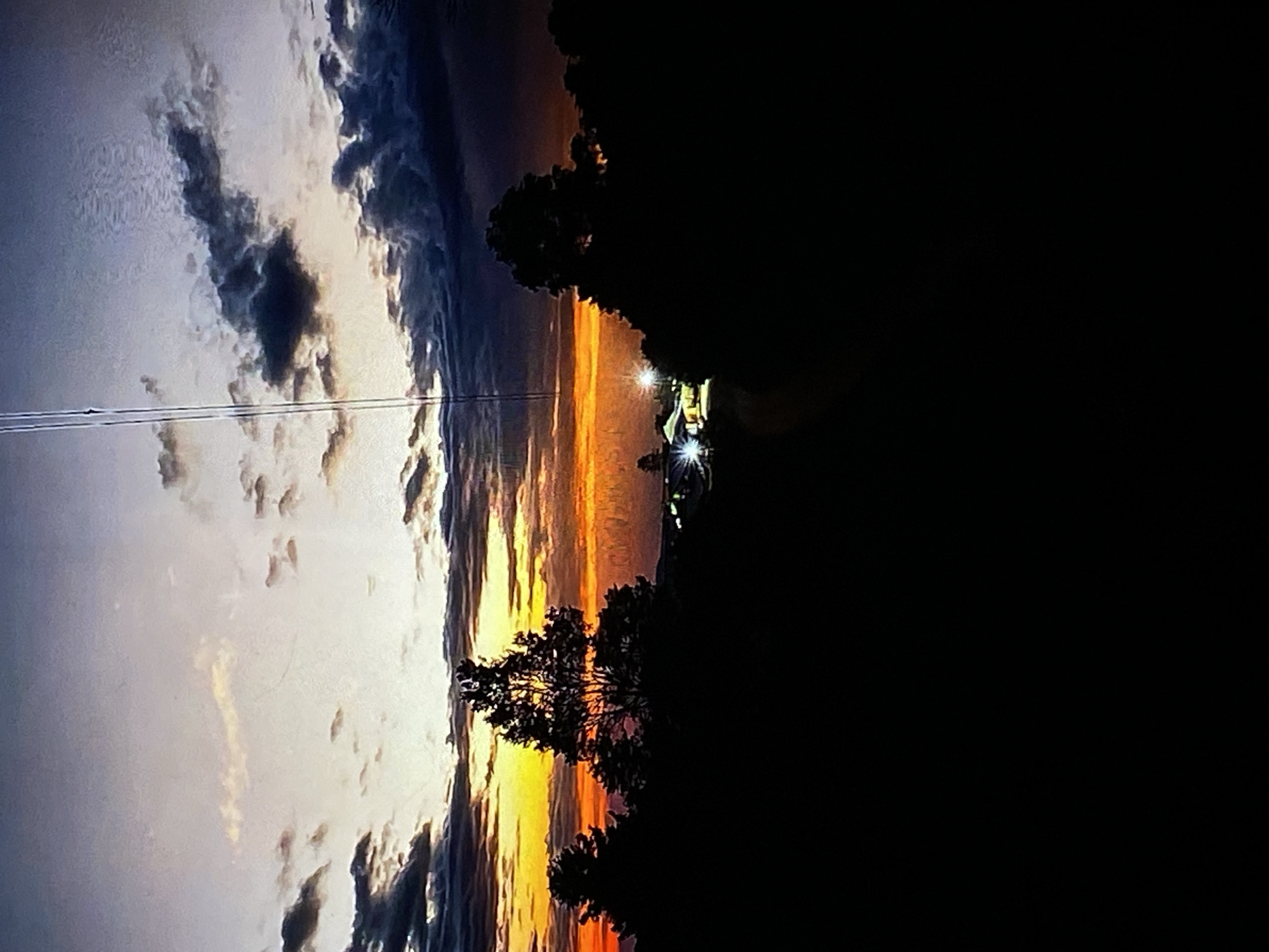 The image captures a sunset over a semi-rural area, possibly on the outskirts of Tinaquillo, Venezuela. The sky is a dramatic display of colors, transitioning from bright yellow and orange near the horizon to darker shades of purple and gray towards the top. Clouds are scattered throughout the sky, some illuminated by the setting sun, while others are dark and ominous.
In the foreground, silhouetted trees create a dark, jagged line against the vibrant sky. These trees appear to be native to the region. In the middle ground, a few buildings are visible, illuminated by artificial lights, suggesting a small settlement or cluster of houses. A tall, slender structure, possibly a communication tower or a power line pylon, stands prominently against the sky, a stark vertical element amidst the natural and man-made landscape. The overall impression is one of a quiet, tranquil evening, with nature's beauty taking center stage, punctuated by the subtle presence of human habitation. The image seems to have been taken from a slightly elevated position, offering a panoramic view of the scene.