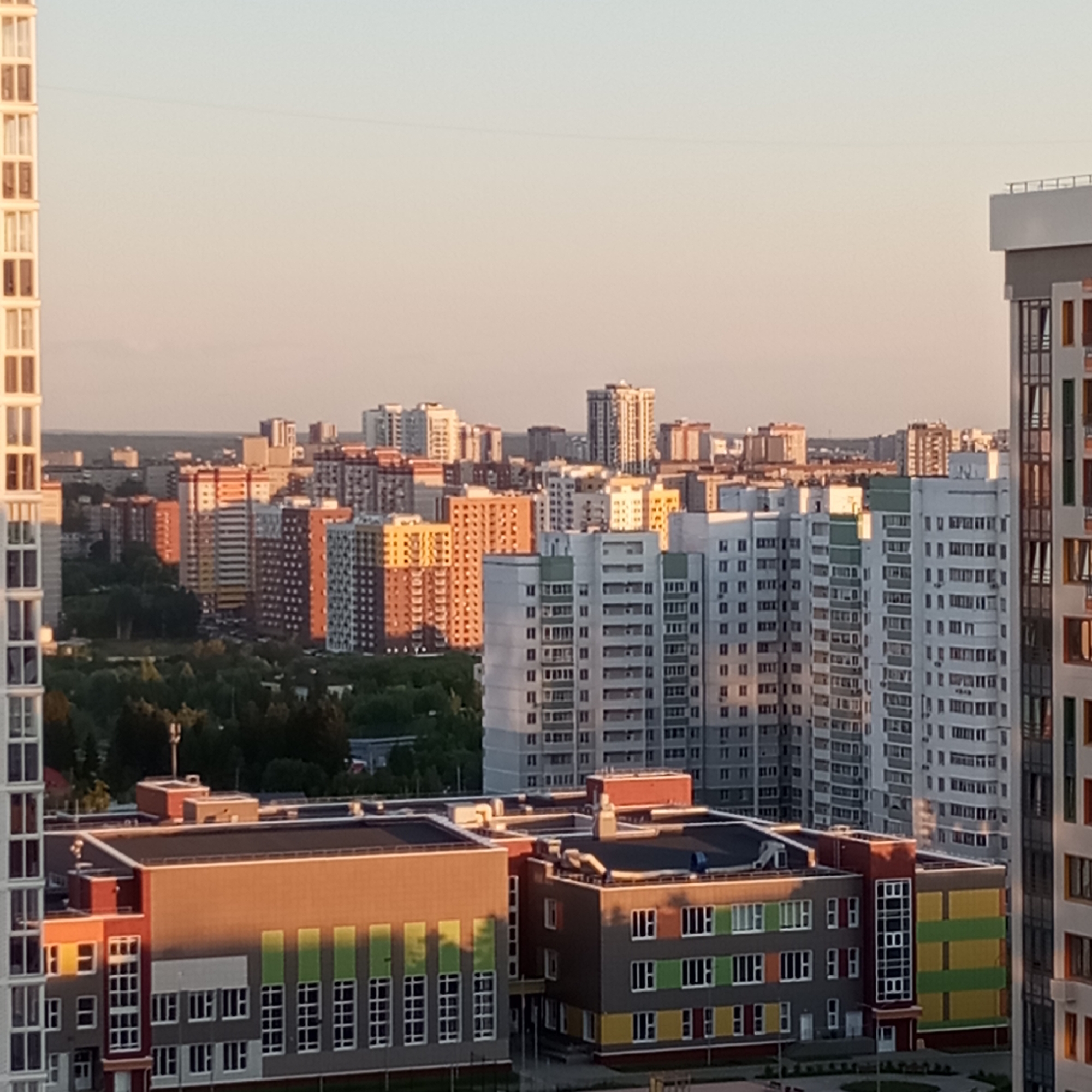 The image presents a panoramic aerial view of a modern urban landscape in Izhevsk, Russia, captured during either late afternoon or early morning, indicated by the warm, directional light and long shadows. The sky is clear, displaying a gradient from pale blue at the zenith to a soft orange-pink closer to the horizon. Faint, thin lines, possibly utility wires, traverse the upper sky.
The midground is dominated by numerous multi-story residential buildings, exhibiting various contemporary architectural styles. Their facades feature a mix of white, grey, beige, brown, and terracotta hues, with many west-facing surfaces illuminated by the warm, golden sunlight. Clusters of green foliage, suggestive of parks or landscaped areas, are interspersed among the distant buildings.
In the foreground, a tall, white residential building with regularly spaced windows is partially visible on the far left. On the far right, another modern, multi-colored high-rise building, partially cropped, features sections of grey, white, orange, and green paneling.
Centrally, two prominent, multi-story buildings with flat roofs, indicative of schools or public facilities, are bathed in the golden light. The left building has a dark grey facade with sections of red/maroon paneling and prominent vertical green accents above windows on its lower section. An antenna-like structure is visible on its roof. The adjacent building to its right features a distinctive facade with horizontal bands of yellow and green paneling, interspersed with windows. Both buildings have flat roofs equipped with visible ventilation or HVAC units. The ground immediately surrounding these foreground structures appears paved and features some green landscaping. No individuals are discernible within the frame.