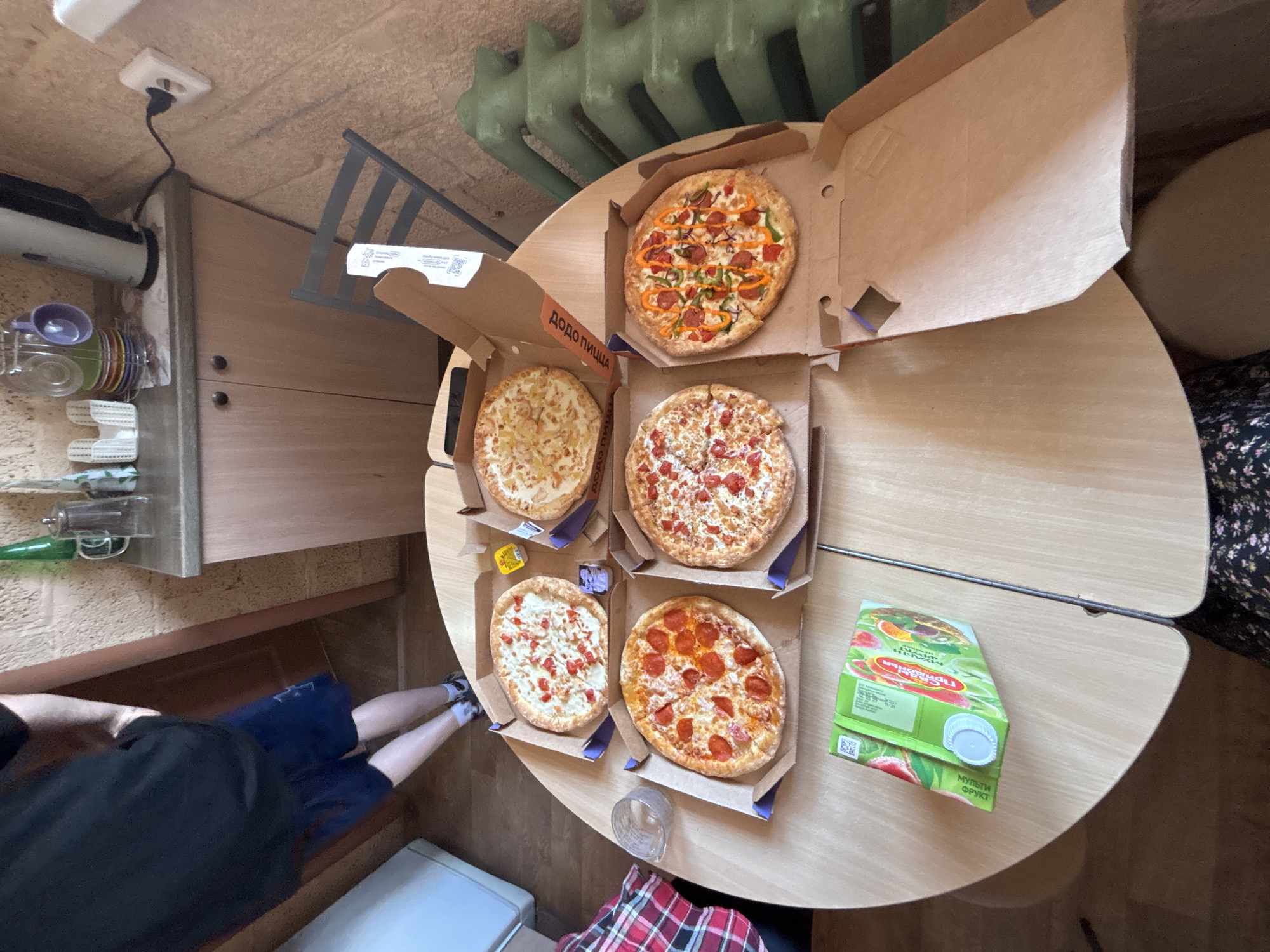 The image shows a table with four open pizza boxes, each containing a different pizza. There is also a carton of juice on the table and a drinking glass. A person wearing a black shirt and blue shorts is partially visible in the lower left corner of the image. A person wearing a red and black checkered shirt is also partially visible in the lower center of the image. The pizza boxes are labeled "Dodo Pizza". Based on this, the location is Русский Вожой, Russia, where Dodo Pizza has locations.