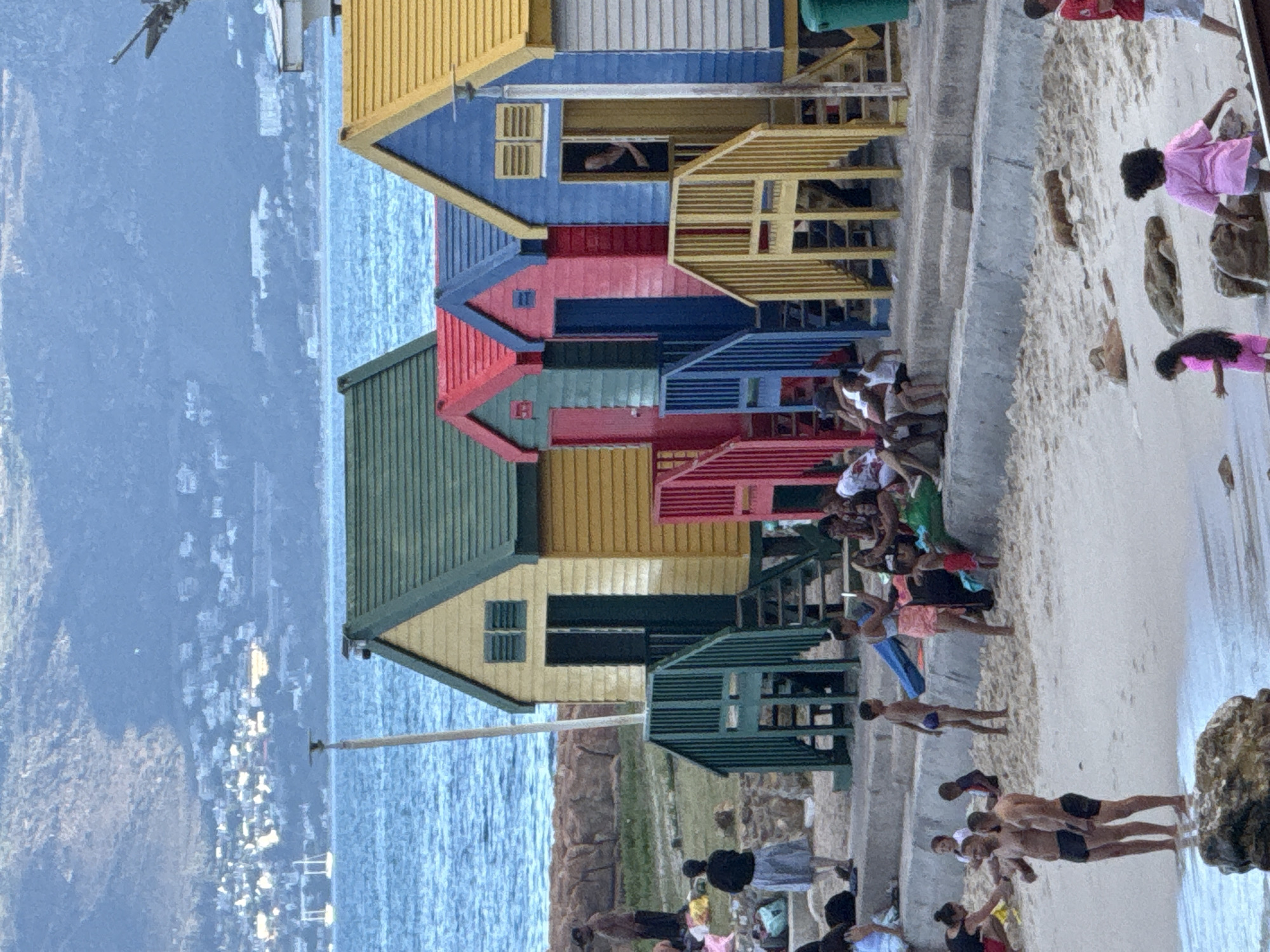 The image shows a row of colorful, wooden beach huts in Muizenberg, a suburb of Cape Town, South Africa, situated along the False Bay coast. The huts are painted in vibrant colors, including yellow, blue, red, green, and orange. Many people are around the huts and on the sandy beach. Some appear to be swimming or wading in the ocean, others are walking or sitting on the sand. The ocean stretches towards the horizon, and a mountain range is visible in the distance. There is a pole perpendicular to the huts going over the beach and into the water.