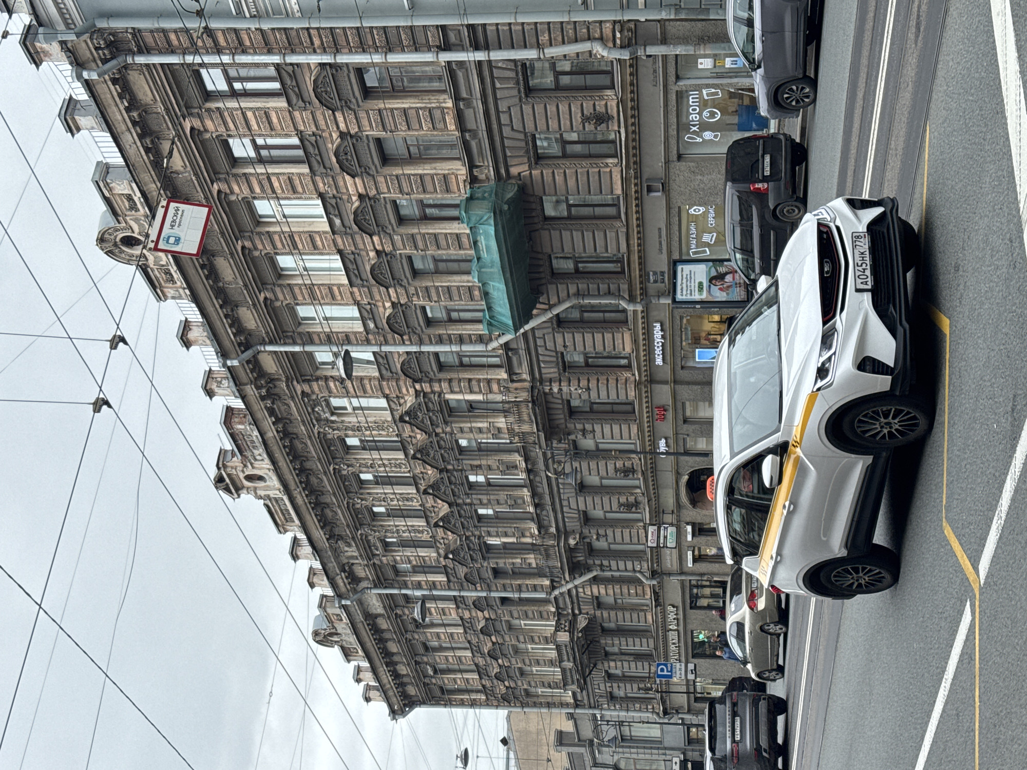 Here is a description of the image:
The image captures a street scene in Saint Petersburg, Russia. Prominently displayed is a white car with a yellow stripe, indicating it is likely a taxi. Its license plate reads "A045HK 778." The car is parked on a street lane marked with yellow lines. Other cars, including gray and black vehicles, are parked behind it.
The backdrop features a large, ornately designed building with numerous windows and architectural details. Several signs are visible on the building, including ones for "Xiaomi," "ЕРТОРСКИЙ ФАРФОР" (Imperial Porcelain), "аксессуары" (accessories), and "обувь." There is also a sign indicating "НЕВСКИЙ проспект" (Nevsky Prospect), a major thoroughfare in Saint Petersburg.
Power lines are visible in the sky above the street. The sky is overcast, contributing to a slightly muted color palette in the image.