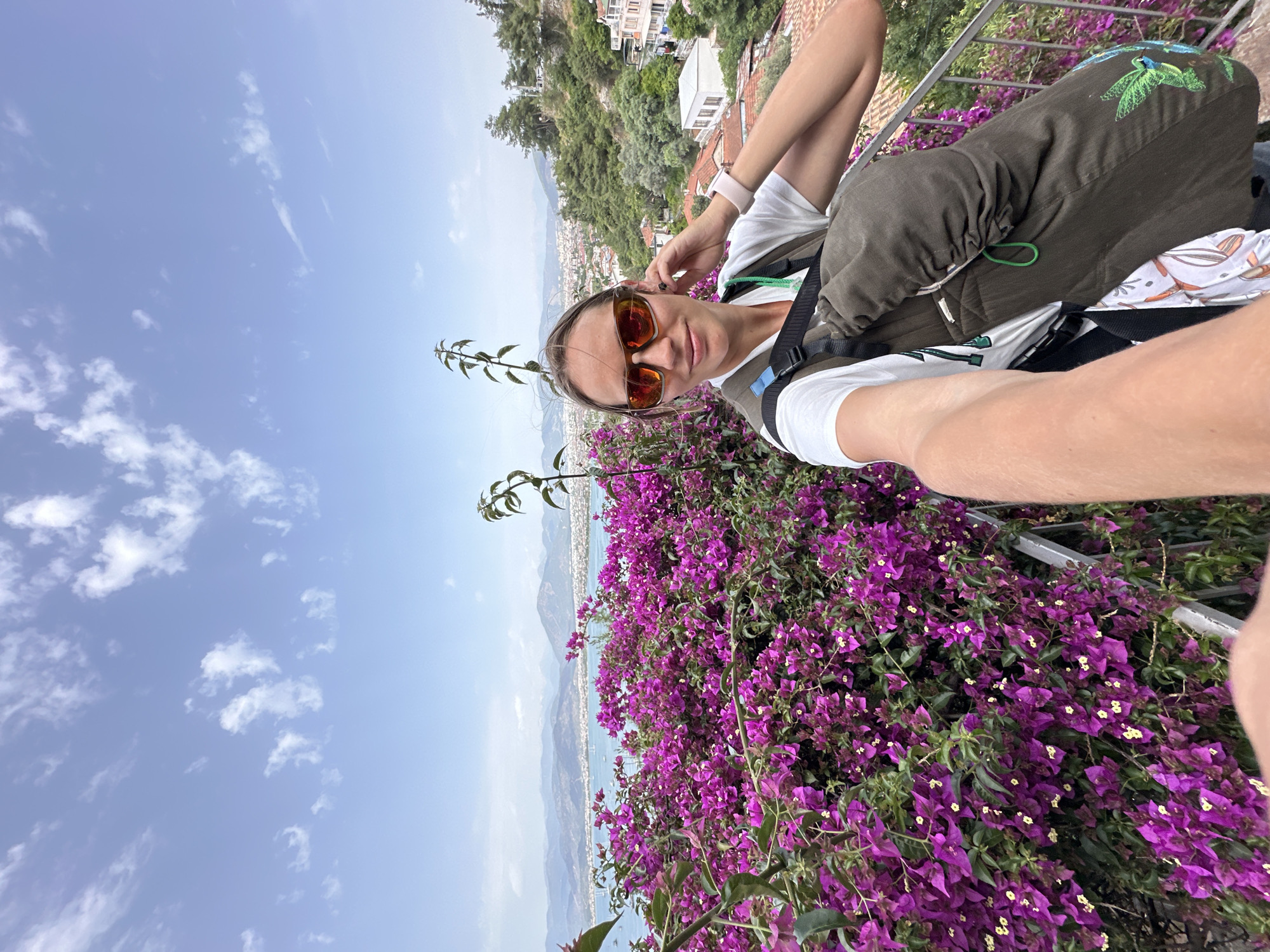 A woman takes a selfie, wearing sunglasses and a backpack, with bougainvillea flowers in the foreground. In the background, the city of Fethiye, Turkey, is visible along the coastline. The sky is blue with scattered clouds.