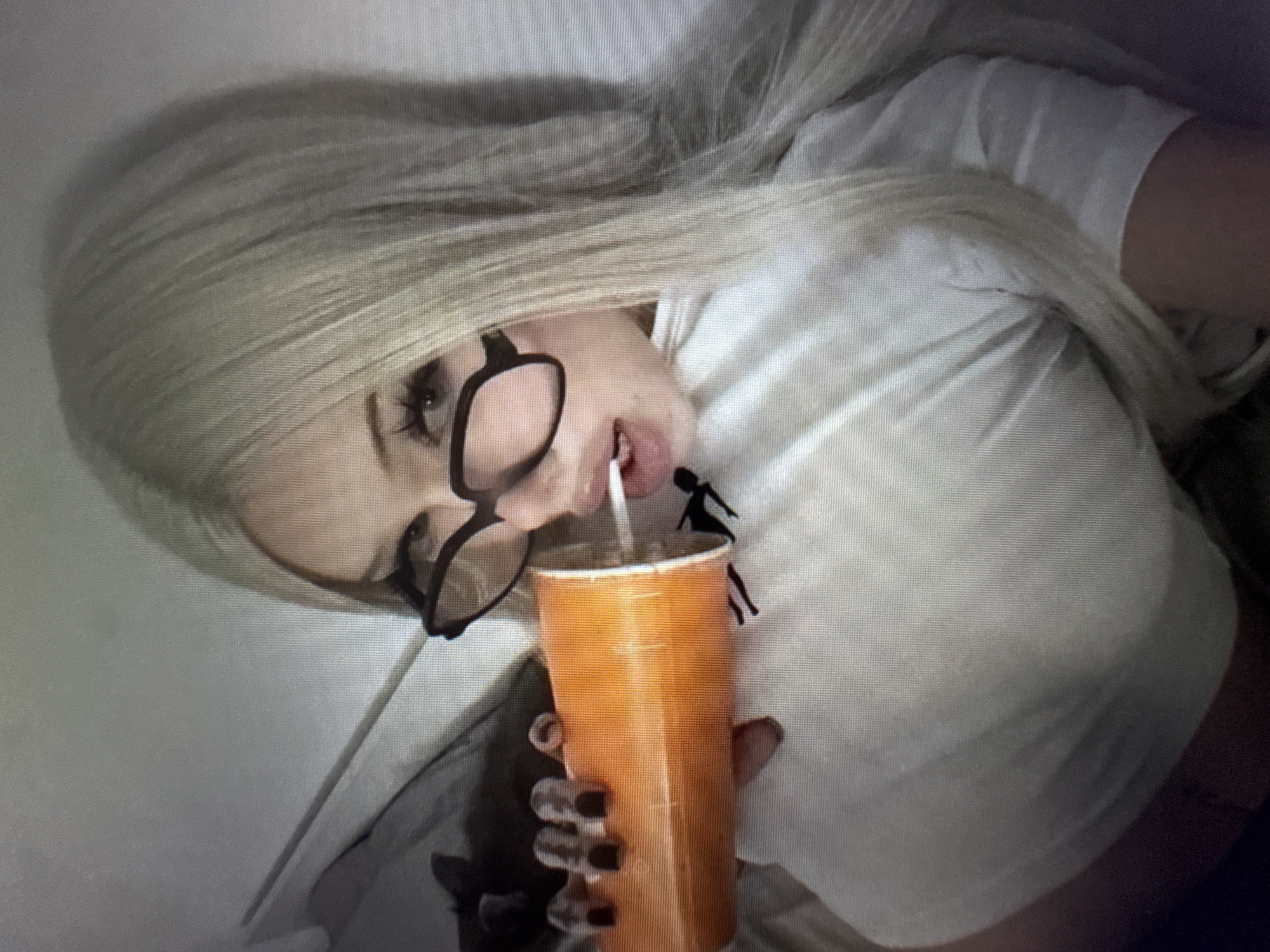 A close-up shot features a person with long, straight, light blonde hair, wearing dark-rimmed glasses and a white short-sleeved t-shirt. The person is positioned with their head tilted, looking towards the viewer. They are holding an orange cup with a white straw and appear to be drinking from it, with their mouth slightly open. Dark nail polish is visible on the fingers holding the cup. On the white t-shirt, near the right shoulder, there is a small black silhouette design of a figure, possibly a person, in a dynamic pose. The background is a soft, light, uniform surface, possibly bedding or a wall. The overall image has a somewhat grainy or pixelated quality. No discernible text is visible.