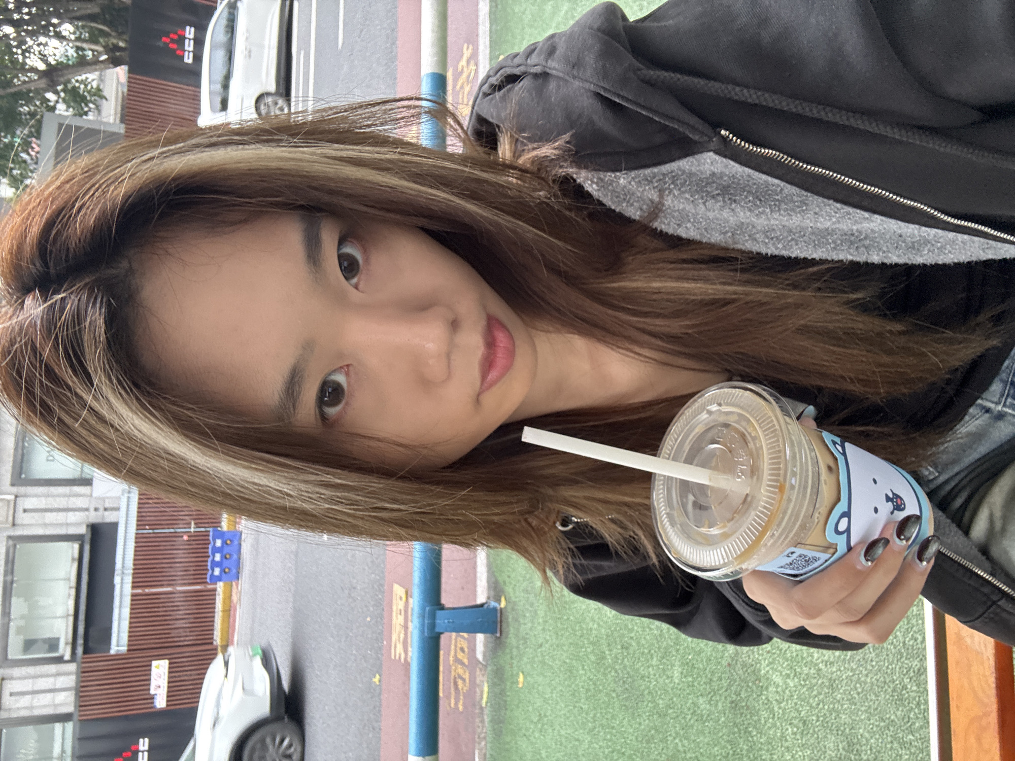A young woman poses for a selfie outdoors, holding a clear plastic cup with a straw. She has long, light brown hair and is wearing a black hooded jacket. The cup is decorated with a cartoon bear design. The background suggests an urban environment with buildings, cars, and a sidewalk. The location is Shapingba District, China. The woman is looking at the camera.