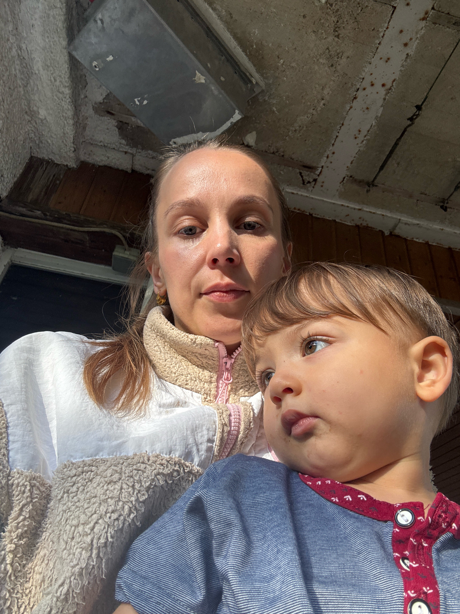 A candid, high-angle medium shot captures a woman and a young child in a close-up selfie. The woman, with fair skin and light brown hair pulled back, is positioned behind and slightly to the left of the child. She is wearing a light-colored zip-up jacket with a beige fleece collar and a white outer shell, and a pair of gold hoop earrings. The child, with light brown hair, is in the foreground, turned to the right and looking off-camera, with a slight pout. The child is wearing a blue striped shirt with a maroon trim and white buttons. The background appears to be an outdoor or semi-enclosed space with a rough, weathered ceiling composed of concrete and exposed beams, and wooden paneling on the walls. The lighting suggests it is daytime, with sunlight casting shadows. The overall setting hints at a rustic or utilitarian area, possibly near a building. The image is undated and the specific location within Überherrn, Germany cannot be determined from the visual information provided.