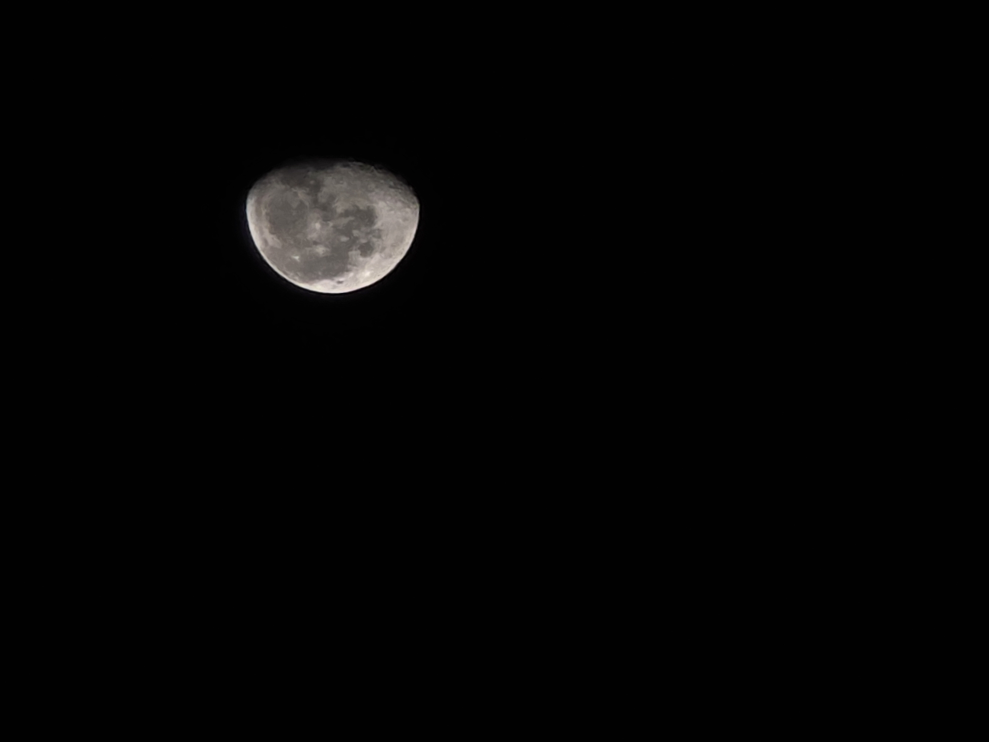 A partially illuminated moon is visible against a black sky. The moon is in the upper right quadrant of the image, and approximately two-thirds of its surface is lit. The surface of the moon displays visible craters and other geological features. The background is entirely black, suggesting a nighttime setting. There are no other discernible objects or subjects in the image.