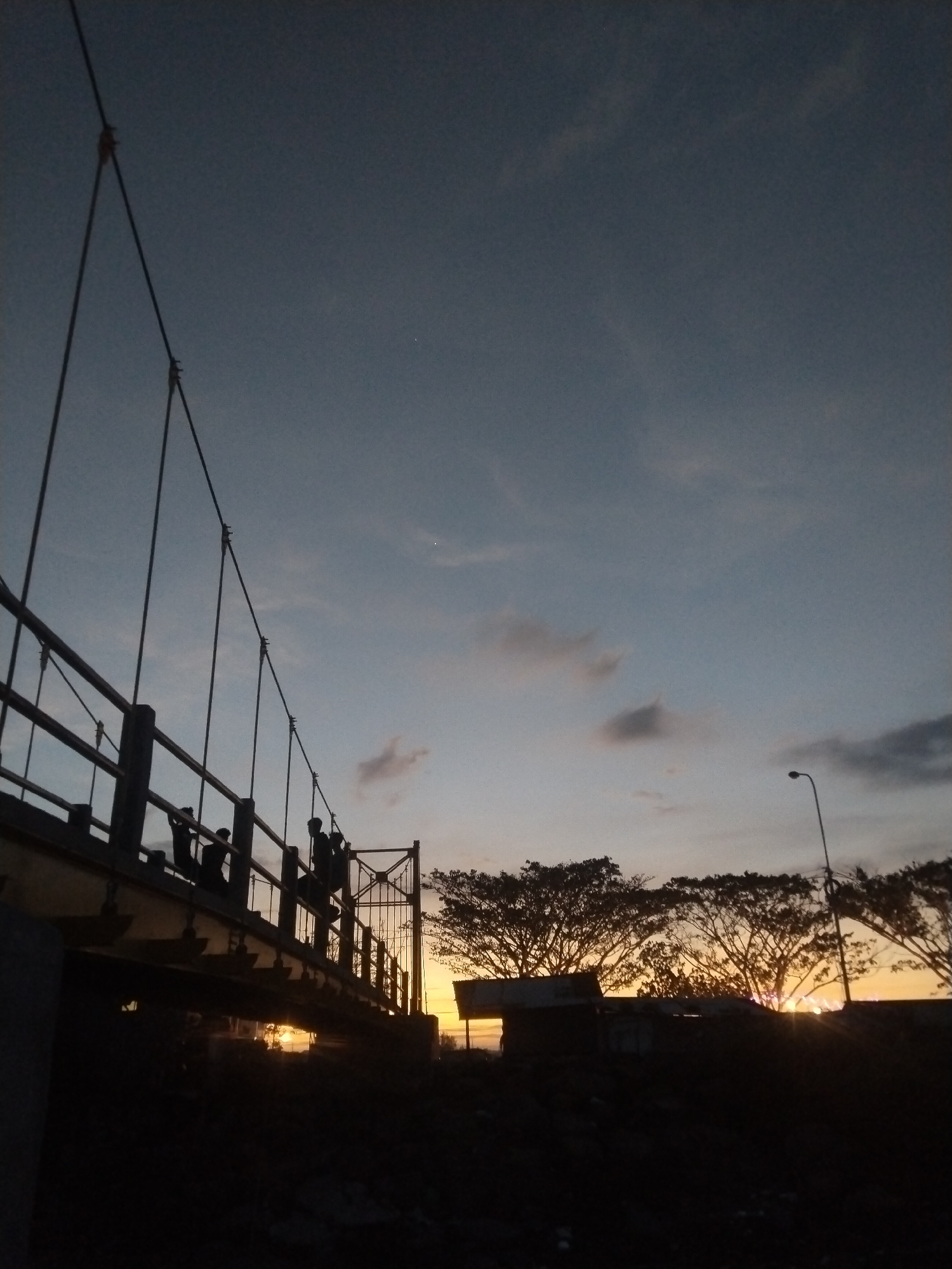 The image captures a suspension bridge in Lhokseumawe, Indonesia, at dusk or dawn. The bridge is silhouetted against the sky, with several people standing on it. The sky is a mix of blue and orange hues, suggesting a sunrise or sunset. Trees and a building are visible in the background, also in silhouette. A streetlight stands to the right of the frame. The overall scene is dark, with the focus on the bridge and the sky.