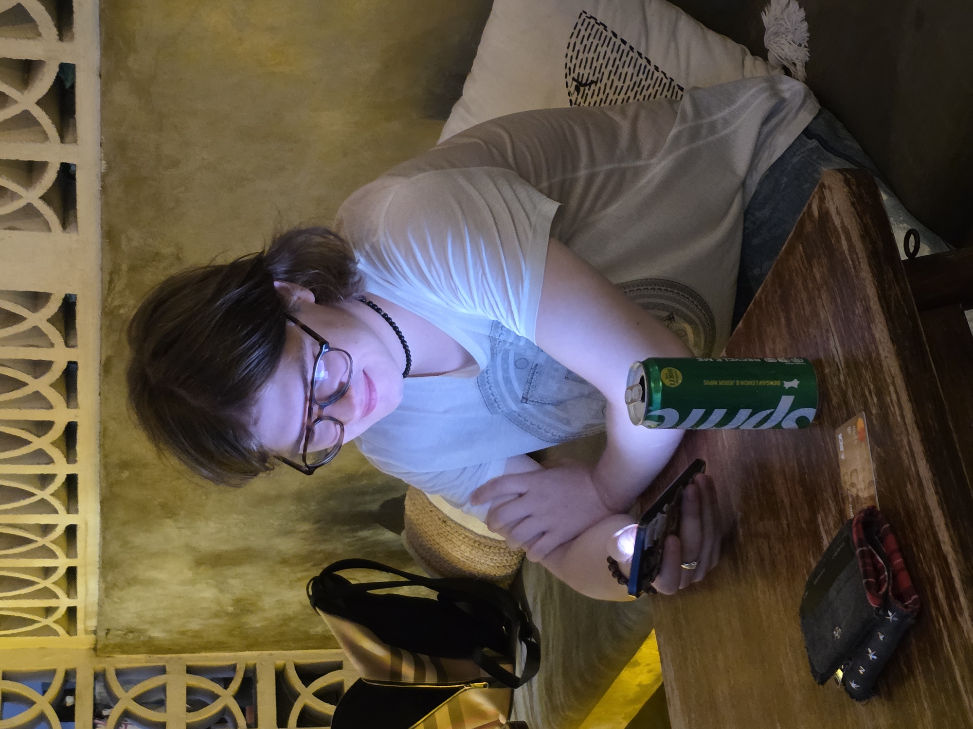 A person with glasses and dark hair pulled back is sitting at a wooden table, looking at their phone. They are wearing a light-colored t-shirt and dark bottoms. To their right, a green can of Sprite is standing upright on the table. A wallet and a card are also on the table in front of the person. In the background, there is a textured wall with decorative elements and a pillow with a black pattern. A yellow bag and a backpack are visible to the left of the person. The scene appears to be in a cafe or restaurant.