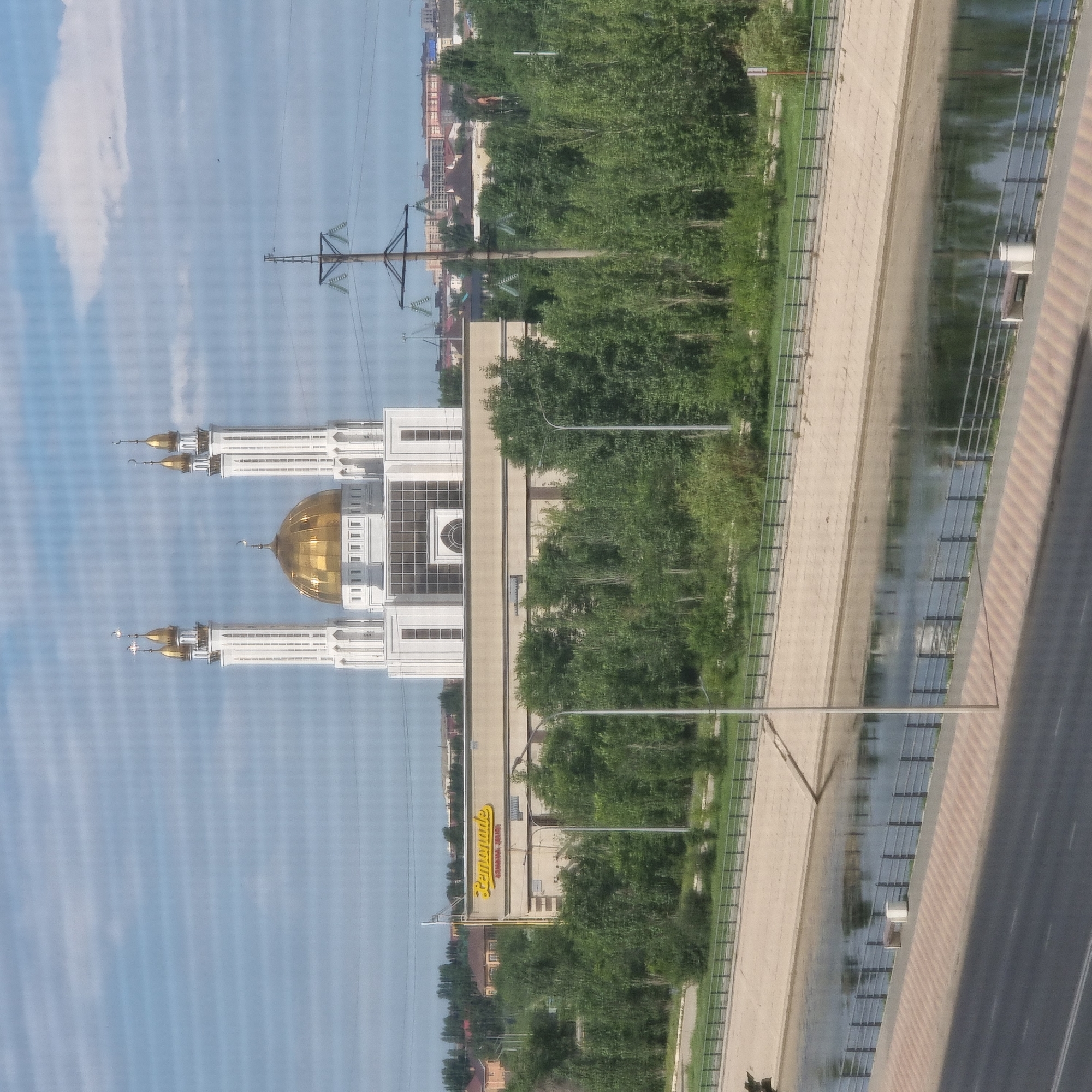 The image depicts a cityscape featuring a prominent mosque with a golden dome and two minarets, situated behind a commercial building with a "Lemonade" sign. The mosque appears to be the Nur-Gasyr Mosque in Aktobe, Kazakhstan, given its architectural style and the surrounding urban landscape. The scene also includes a river or canal in the foreground, bordered by concrete embankments and a pedestrian walkway with a metal railing. Trees and other buildings are visible in the background, along with power lines and a utility pole. The sky is partly cloudy, suggesting daytime. No people are clearly visible in the image.