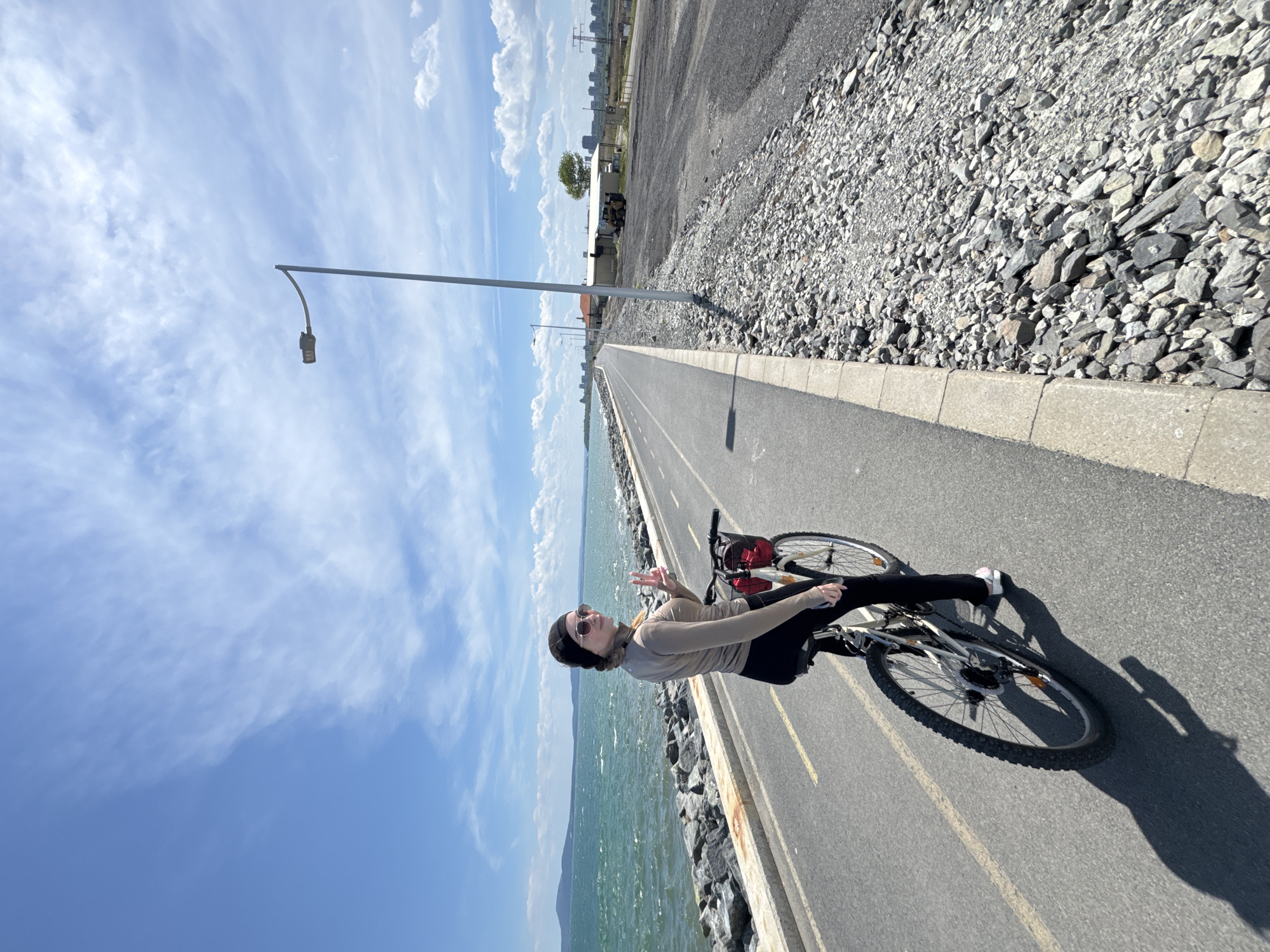 A cyclist pauses on a paved seaside path in Burgas, Bulgaria, under a partly cloudy sky. The path, bordered by the sea on one side and a rocky, gravel area on the other, stretches into the distance, flanked by a light pole. In the background, a cityscape is visible. The person is facing the camera, holding up their fingers in a peace sign, and wearing sunglasses, a head covering, a light-colored long-sleeved shirt, and dark pants. They are seated on a bicycle, with a red basket behind the seat. Their shadow extends across the path. The sea, on the left, is choppy.