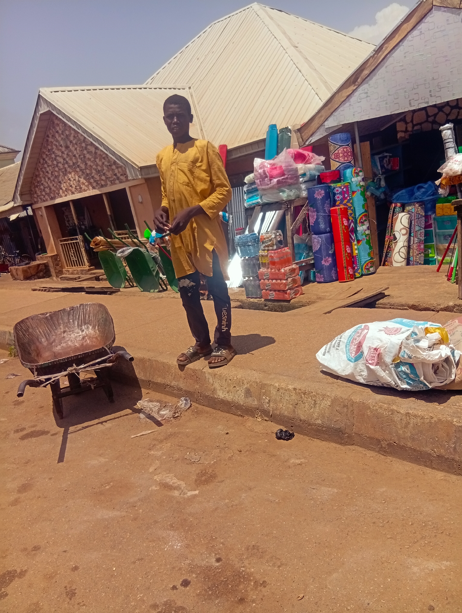 A young man stands in front of a market stall in Bukuru, Nigeria. He is wearing a yellow, long-sleeved tunic and dark pants with white text on the right leg. He is holding a small item in his hands. The market stall behind him is stocked with various goods, including what appear to be rolled-up mats, bags of products, and stacks of bottled water. In the foreground, to the left, is a metal wheelbarrow. To the right, on the ground, is a large, white, plastic-wrapped bag, possibly containing goods or supplies, with blue and red text visible. The ground is a dusty, reddish-brown dirt, and a concrete curb runs along the front of the market. Several other wheelbarrows are visible further back, lined up against the stall. The scene appears to be set during the day, with bright sunlight casting shadows. The sky is a pale blue with some scattered clouds. The overall impression is of a busy marketplace.