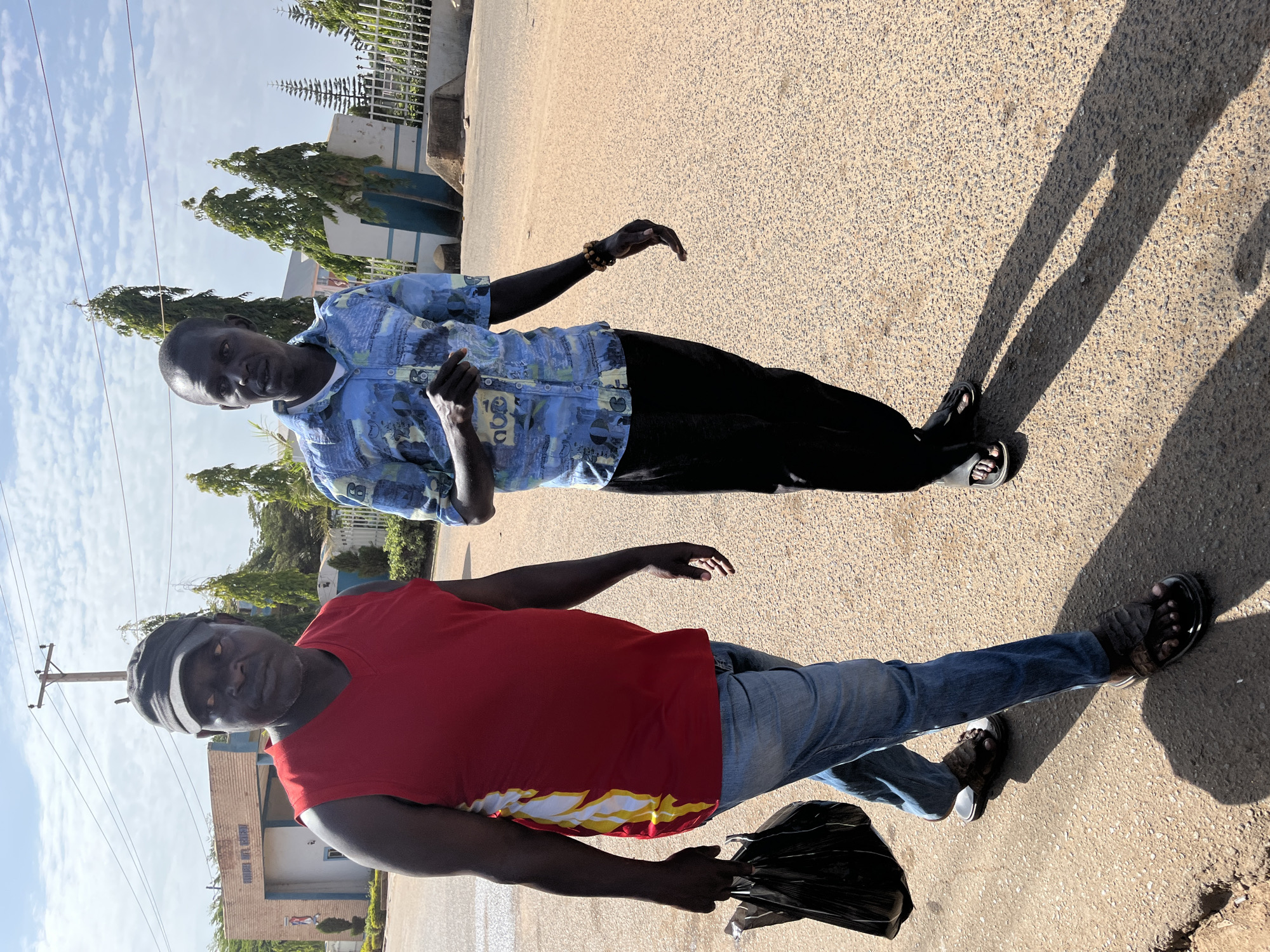 Two dark-skinned men stand on an asphalt road. The man in the foreground wears a red t-shirt, blue jeans, a black cap, and sandals. He holds a black bag. The man in the background wears a blue patterned shirt, black pants, and sandals. They both appear to be walking or posing for a photo. The setting appears to be an outdoor environment in Jos, Nigeria. A building, trees, and a cloudy sky are visible in the background.
