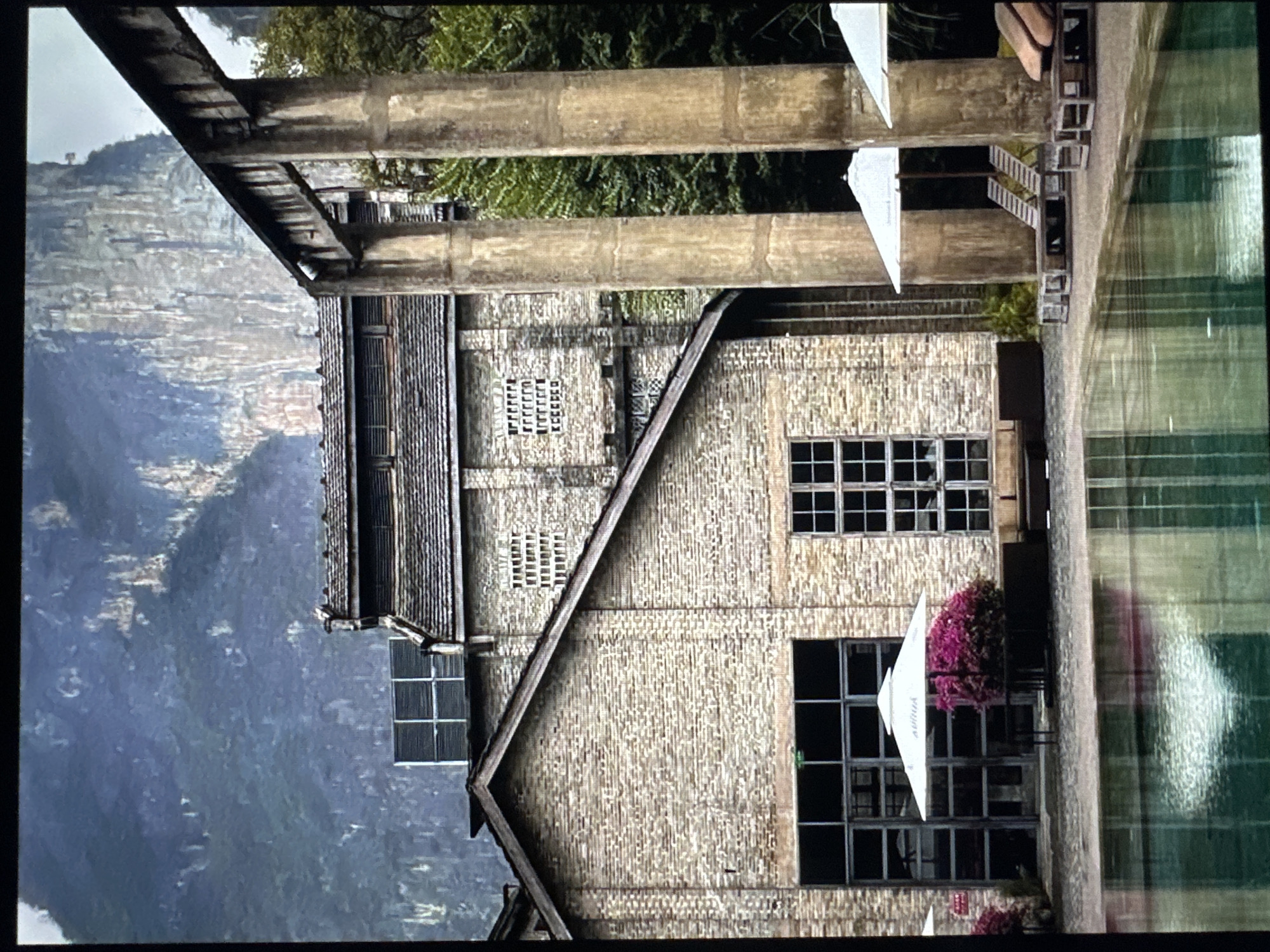 This media file captures a building with a rustic, industrial aesthetic, possibly an old factory or warehouse, nestled against a dramatic mountainous landscape. The building's exterior is constructed with rough-hewn stone or brick, featuring large, multi-paned windows and a pitched, weathered wooden roofline. Above the main structure, massive concrete beams suggest former industrial supports or a partially demolished upper level.
In the foreground to the right, a shimmering green surface, likely a pool of water or a reflective floor, is visible. White umbrellas and vibrant pink flowering bushes add touches of color and suggest an outdoor seating or leisure area. The reflection in the green surface hints at the presence of the building and possibly the sky.
The background is dominated by a steep, rocky mountain face with a soft, hazy atmosphere, indicating a possibly overcast or misty day. The overall mood is one of quiet contemplation, with the juxtaposition of the sturdy, aged architecture and the raw natural beauty of the mountains. The image is oriented vertically, rotated 90 degrees counterclockwise, so the building appears to extend upwards into the mountains. No people are visible, and no text is clearly discernible. The time of day appears to be daylight, given the lighting.