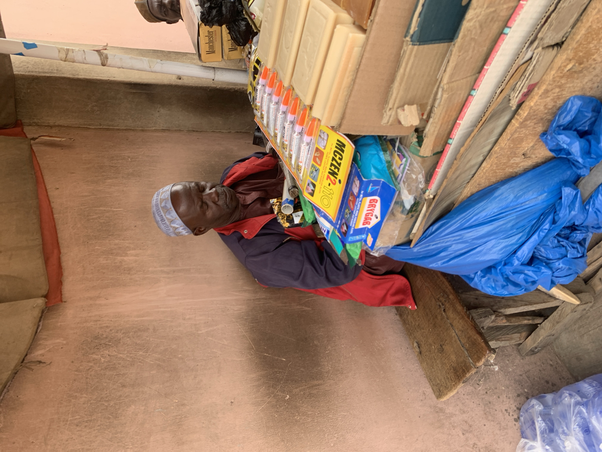 An elderly African man with dark skin, a white and blue patterned kufi cap, wearing a red and blue jacket, sits next to a small, makeshift shop in Jos, Nigeria. The shop is a collection of cardboard boxes, wooden boards, and blue plastic bags containing merchandise like soap, glue, and other small items. The scene appears to be outdoors, with a concrete or tiled floor visible. The lighting suggests it is daytime.