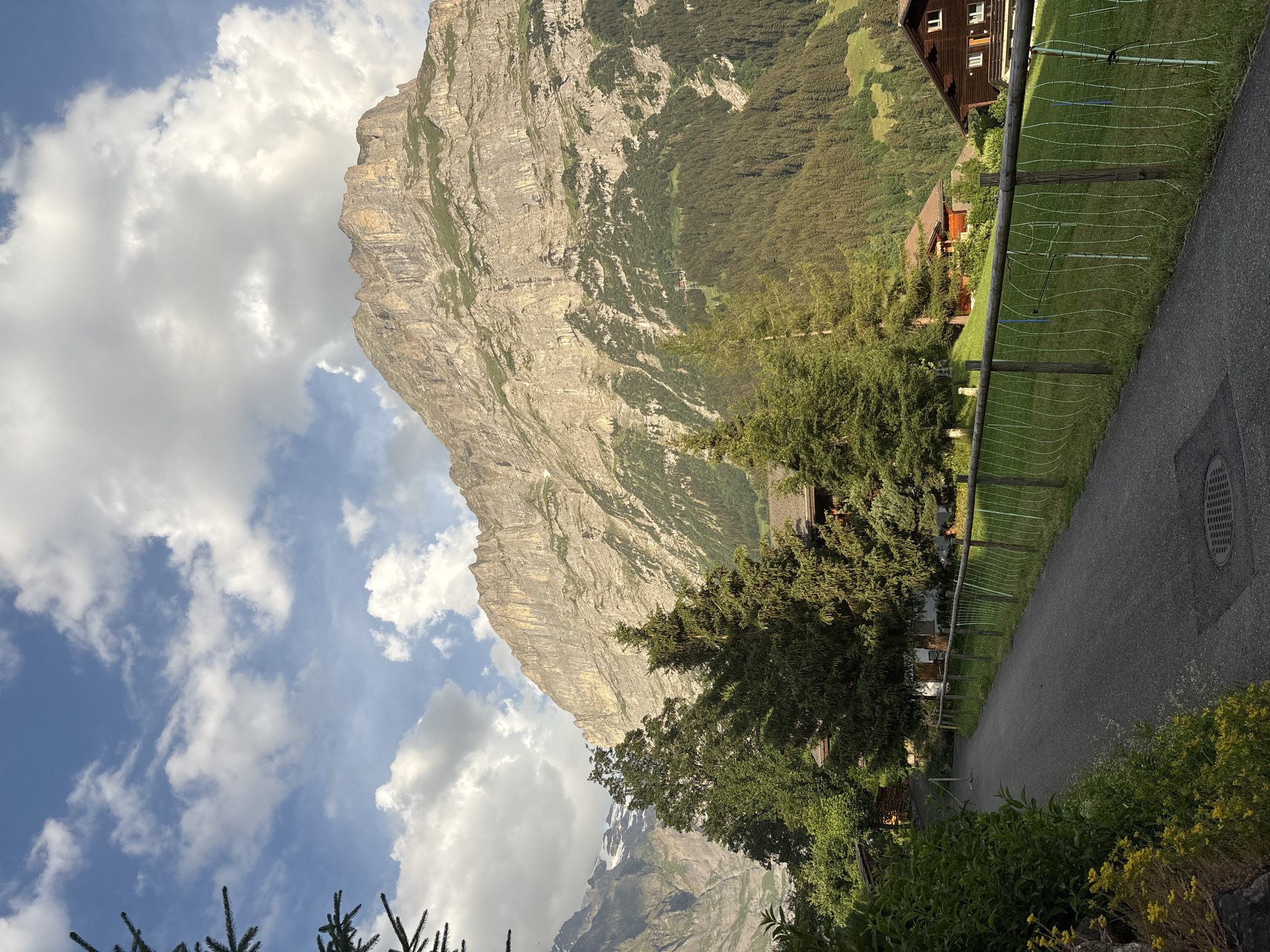This media file captures a scenic view of a mountainous landscape with a village nestled at its base. The dominant subject is a massive, rugged mountain face, characterized by its sheer cliffs and rocky texture, partially covered in sparse vegetation. Below the mountain, a dense green forest blankets the slopes, transitioning into a neatly mown grassy area.
Several human-made structures are visible: a traditional wooden house with a dark brown facade and white trim, and another smaller red-roofed building. A paved path or road curves through the scene, bordered by a wooden fence with white netting. This suggests a residential or tourist area.
The sky is a mix of bright blue and dramatic white clouds, indicating a partly cloudy day. The lighting suggests it is either late afternoon or early morning, with the sun casting a warm glow on the mountain face. The overall mood is peaceful and picturesque, highlighting the grandeur of nature and the harmonious integration of human settlement within it. The location is identified as Grindelwald, Switzerland, which aligns with the alpine scenery. There is no visible text within the image.