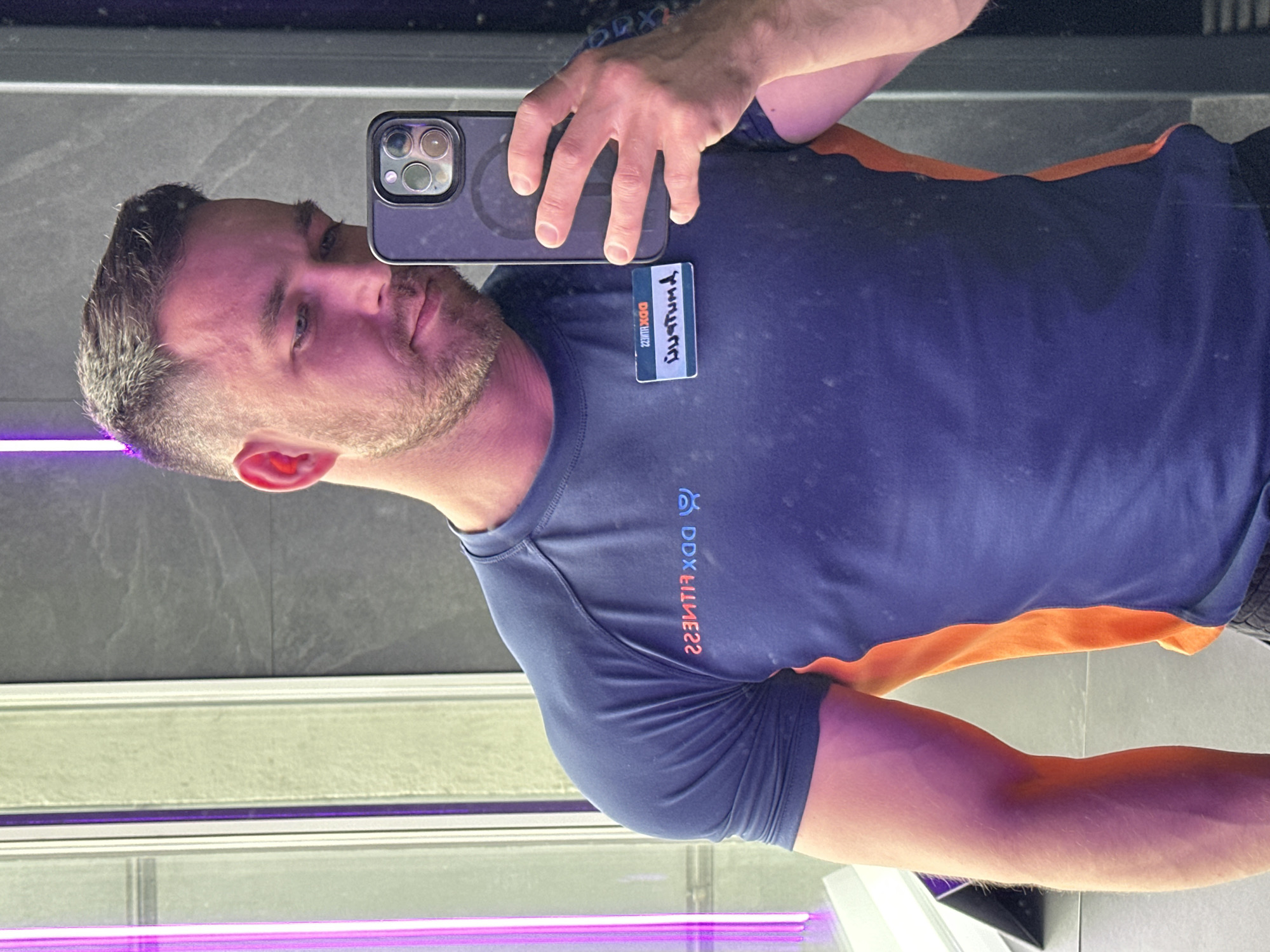 A man is taking a selfie in a gym mirror. He is wearing a blue t-shirt with orange accents and a DDX Fitness logo, along with a name tag with the name "Custom T" handwritten on it. He is holding a smartphone in his right hand to capture the image. The background shows gray tiled walls and a purple neon light. Based on the DDX Fitness logo, the location is likely in Perm, Russia.