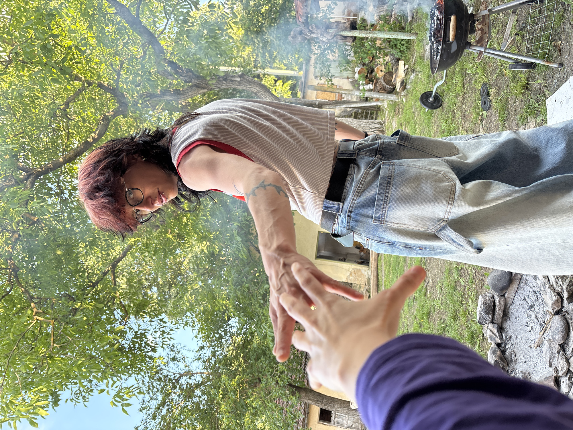 The image depicts an outdoor scene in what appears to be Karlukovo, Bulgaria, characterized by lush green trees and a bright sky. A person with reddish-brown hair and glasses is the primary subject, wearing a striped tank top and baggy blue jeans with a black belt. They are leaning forward, extending their right arm towards the viewer. On their arm, there is a tattoo visible. The perspective is from a lower angle, looking up at the individual, with another person's arm and hand extending towards them, suggesting a potential interaction. A black barbecue grill with smoke emanating from it is present in the background, along with stacked firewood and buildings, indicating a residential or recreational area. A ring of rocks and a small fire pit is visible near the bottom of the frame. The overall atmosphere suggests a casual, outdoor gathering.
