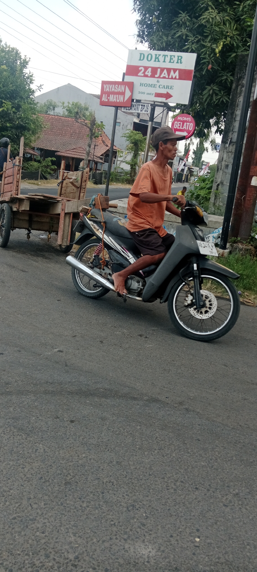 An Indonesian man wearing a dark brown cap and an orange t-shirt rides a black motorcycle pulling a wooden cart. The man is barefoot and is looking forward. The motorcycle has a license plate that reads "AE 2415 RG". The wooden cart is filled with what appears to be agricultural produce, tied with rope. In the background, there is a sign that reads "DOKTER 24 JAM & HOME CARE" and another sign that says "YAYASAN AL-MA'UN". There is also a sign for "MOMS GELATO". The scene appears to be in a rural or semi-urban area, with trees and buildings visible. The road is paved and appears to be a two-lane street. The weather is overcast. The location is identified as Maguwoharjo, Indonesia.