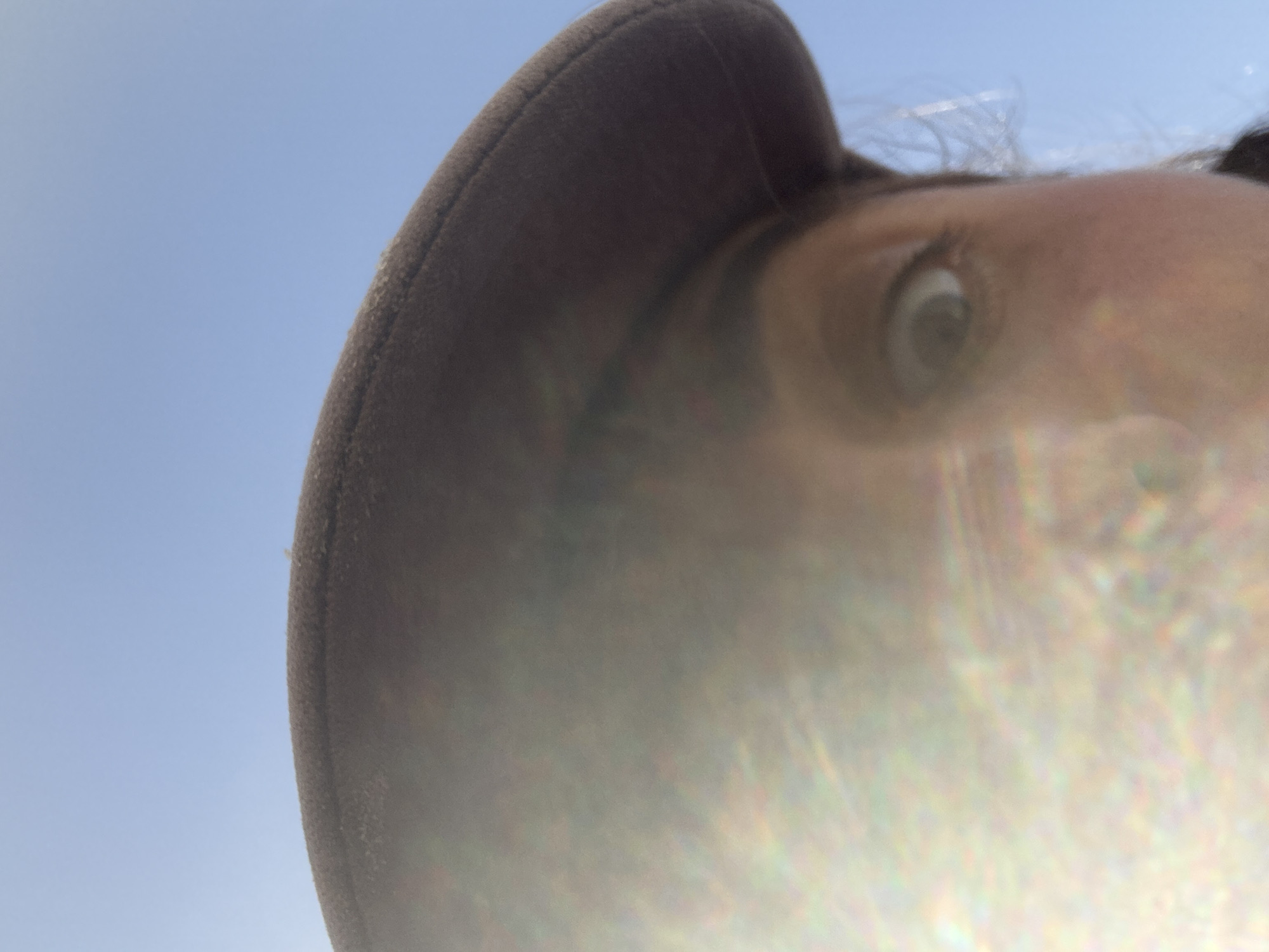 A close-up, low-angle photograph shows a person wearing a hat against a blue sky. Only one eye of the person is clearly visible. The bottom portion of the image is obscured by a bright, indistinct light, possibly due to direct sunlight hitting the camera lens. The scene does not contain any information about the city of Tel-Aviv, Israel.