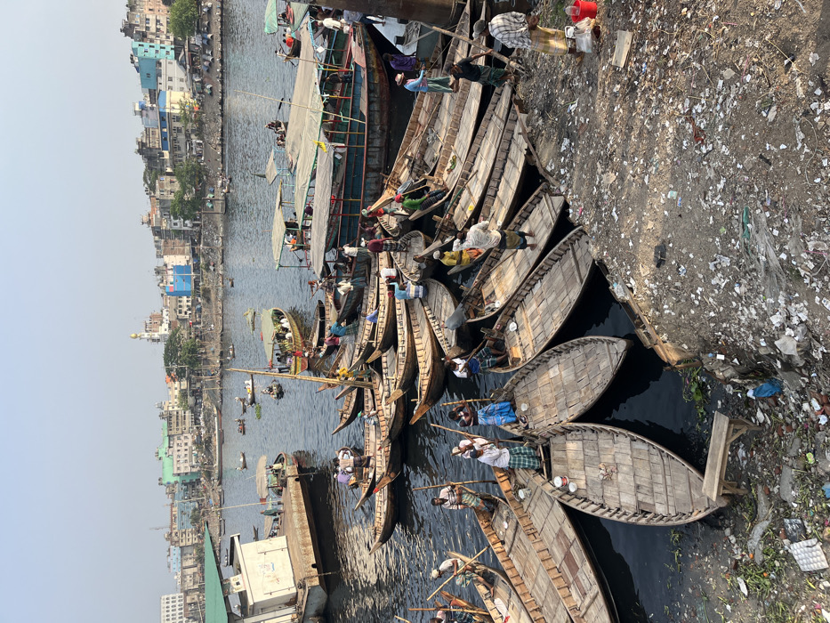 This image depicts a densely populated riverfront scene on the Buriganga River near Sadarghat in Dhaka, Bangladesh. The foreground shows a muddy, unpaved bank heavily littered with various types of refuse, including plastic bags, bottles, paper, and organic debris. A small, light-colored wooden bench is situated on this debris-strewn bank.
The river is actively used, populated by numerous traditional wooden boats. These range from small, open-decked skiffs, many of which are clustered and moored along the immediate bank, to several larger passenger boats with canopy roofs positioned further out in the water. Approximately 20-30 individuals, primarily adult males, are visible throughout the scene. They are engaged in various actions, including poling the small boats, standing within them, or standing on the riverbank. Their attire typically consists of lungis (checked or striped fabric wraps) and shirts. For example, a man in a white and green striped lungi poles a boat in the lower-left, while another in a blue and white striped lungi stands in an adjacent vessel. On the right bank, a man in a plaid shirt and light-colored lungi stands next to two red plastic buckets. The river water appears dark and somewhat murky.
The background reveals a dense urban skyline along the opposite bank, characterized by numerous multi-story buildings of varying colors (blue, green, white, brick) and architectural styles. A prominent white mosque featuring a gold dome and a single minaret stands out among these structures. The sky is clear and light blue, indicating daytime conditions with a visible atmospheric haze over the distant cityscape.