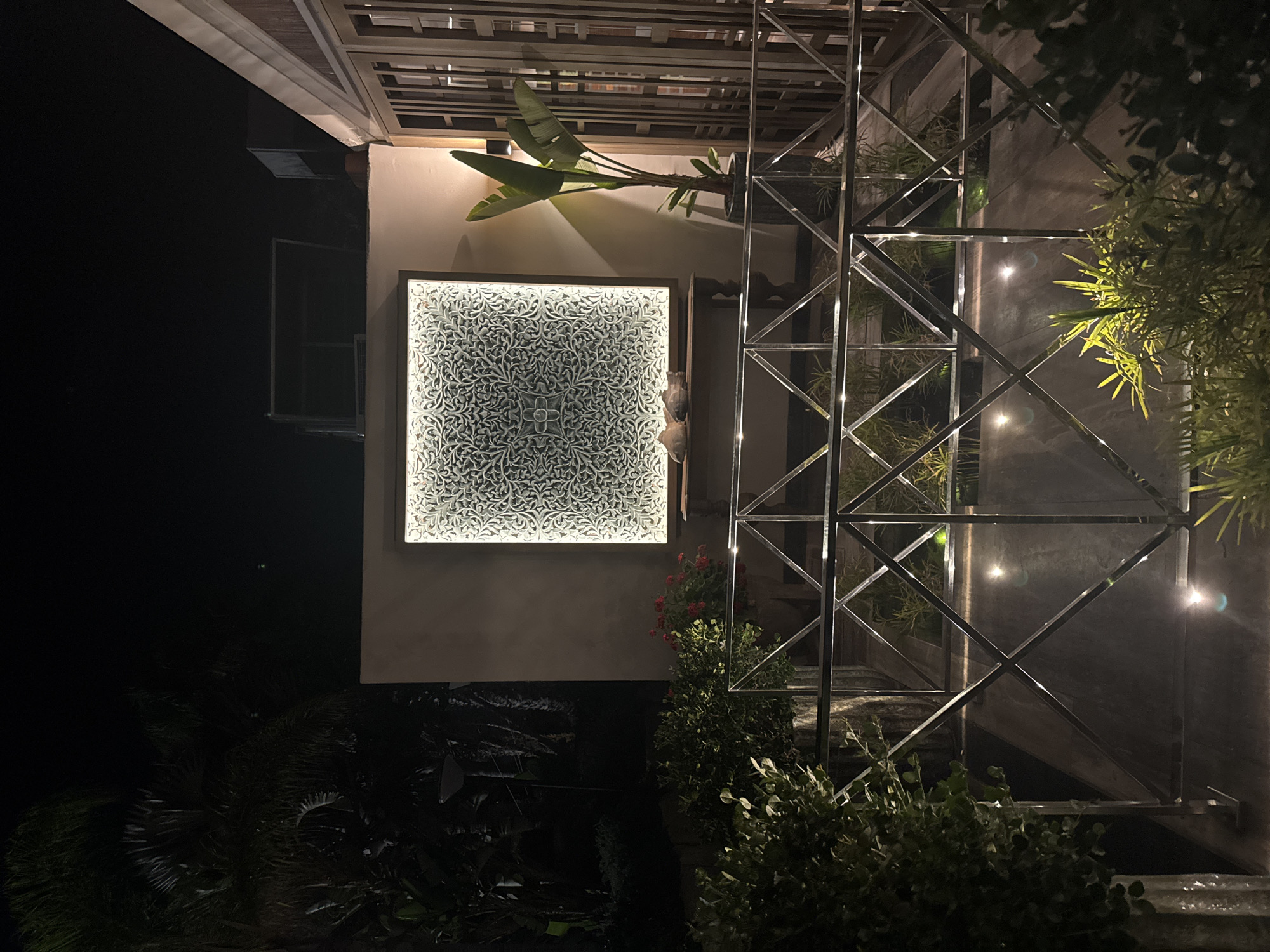 At night, an illuminated square artwork with intricate white patterns is centered on a cream-colored wall. Below the artwork, a dark wooden table displays two decorative fish sculptures. To the right of the artwork, a tall potted plant with large green leaves stands against the wall. A modern building with wooden latticed doors is visible on the far right. In the foreground, a silver metallic railing with a diamond pattern separates the viewer from a garden area. The garden features low-lying plants and subtle ground lighting. Lush, dark greenery and palm fronds are visible in the background on the left, suggesting an outdoor or semi-outdoor setting. The location is identified as Agios Tychona Community, Cyprus.