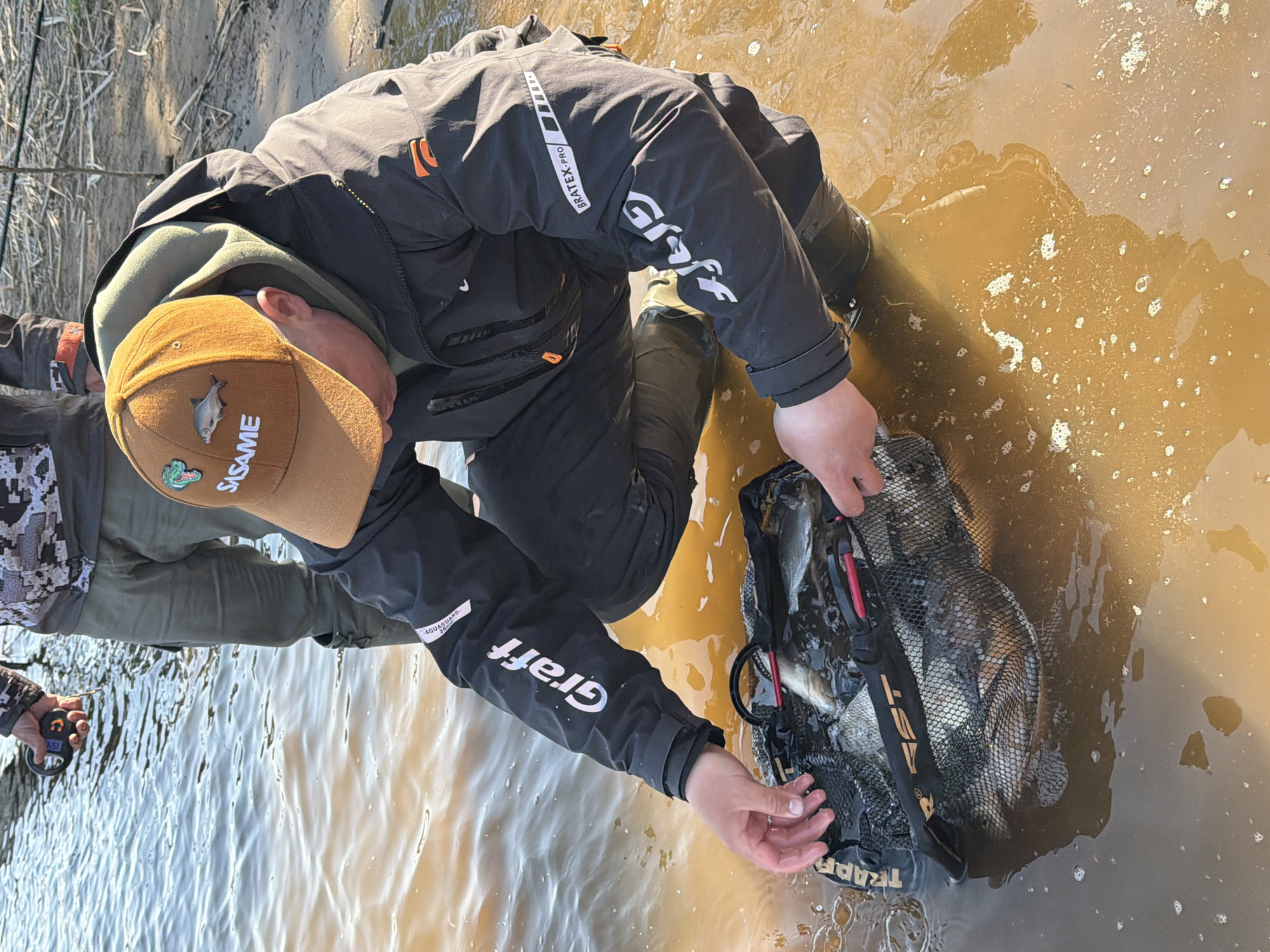 A person wearing Graff-branded clothing is kneeling in a muddy body of water, with their hands near a net containing several fish. A second person is partially visible to the left, holding a device. The water is murky, and the bank of a river or pond is visible in the upper left. The person in the image is wearing a cap with a fish and "SASAME" embroidered on it. The environment suggests an outdoor setting. The clothing is suitable for protection from the weather and elements, and the presence of the net implies an activity like fishing. I cannot confirm the location as Chaykovsky, Russia, without further information.