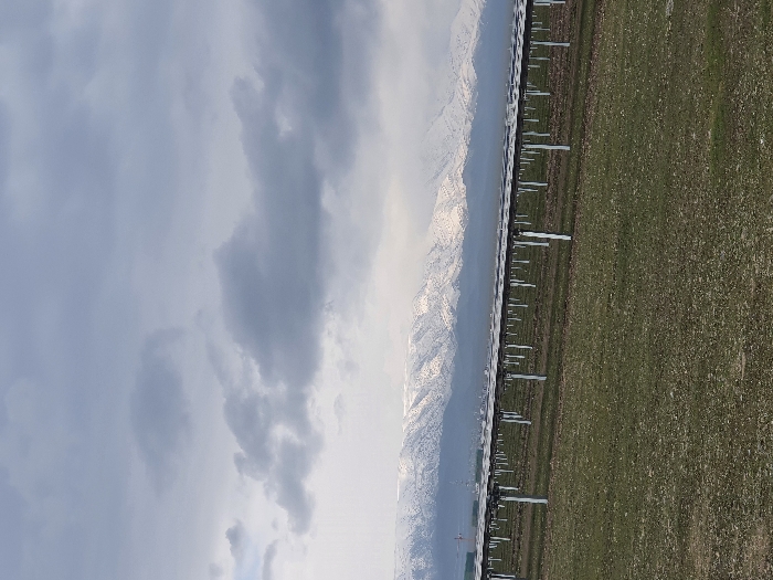 The landscape features a field of green dotted with repetitive white structures, likely solar panels, extending into the distance. Jagged, snow-capped mountains rise in the backdrop, partially obscured by haze. Overcast skies dominate the scene, with dense gray clouds filling the frame.